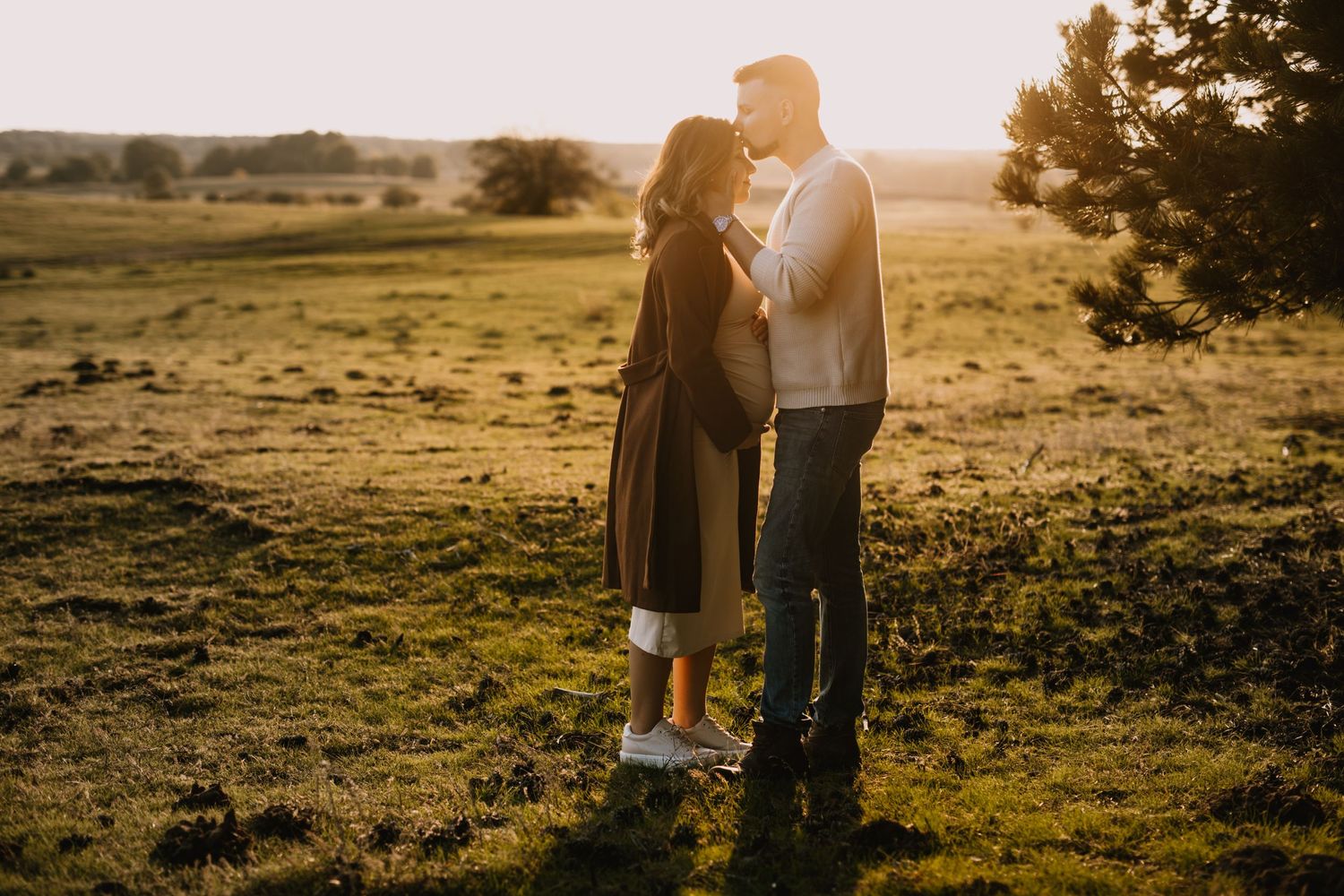 A couple stands close together in a sunlit meadow at golden hour.