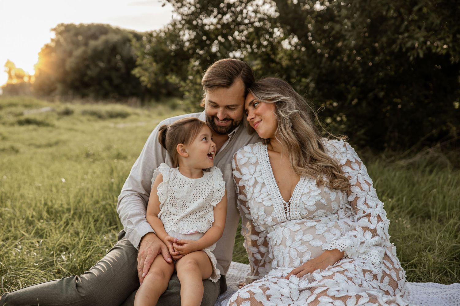 A family enjoys a tender moment together in a sunlit meadow at golden hour wearing white floral outfits.