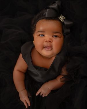 Adorable baby girl in black dress and rhinestone bow headband poses on black fabric backdrop.