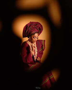 Figure in burgundy traditional attire with matching gele headwrap and beaded necklace captured in dramatic lighting.