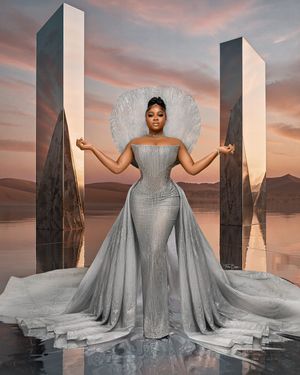 Someone in a flowing silver gown with dramatic cape stands amongst geometric stone pillars at sunset.