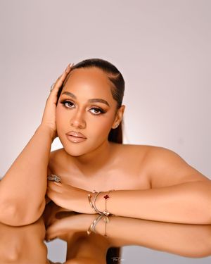 Artistic portrait showing shoulders and hands in a soft, warm-toned studio setting with delicate makeup and jewelry.
