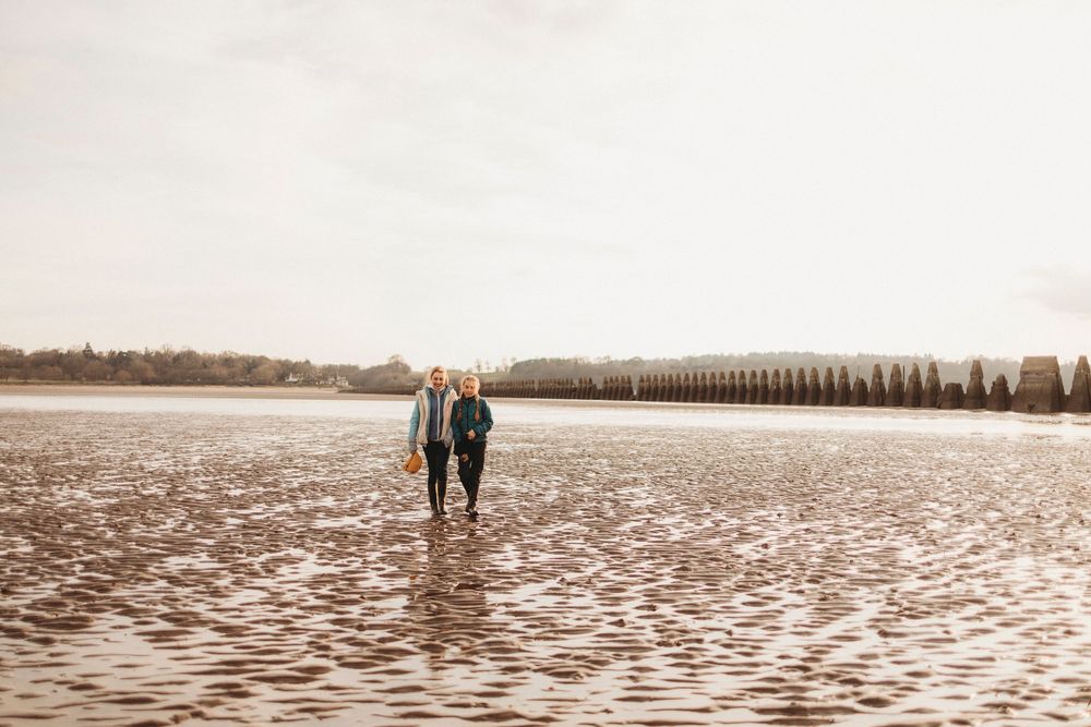 Two people walk hand in hand along a wet beach with rows of trees visible in the distance.