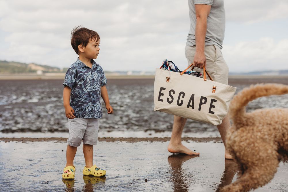A child in striped overalls plays on a beach with a golden retriever dog running alongside them in the sand.