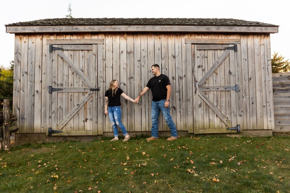 A couple stands in front of a rustic wooden barn during an autumn engagement photo session.