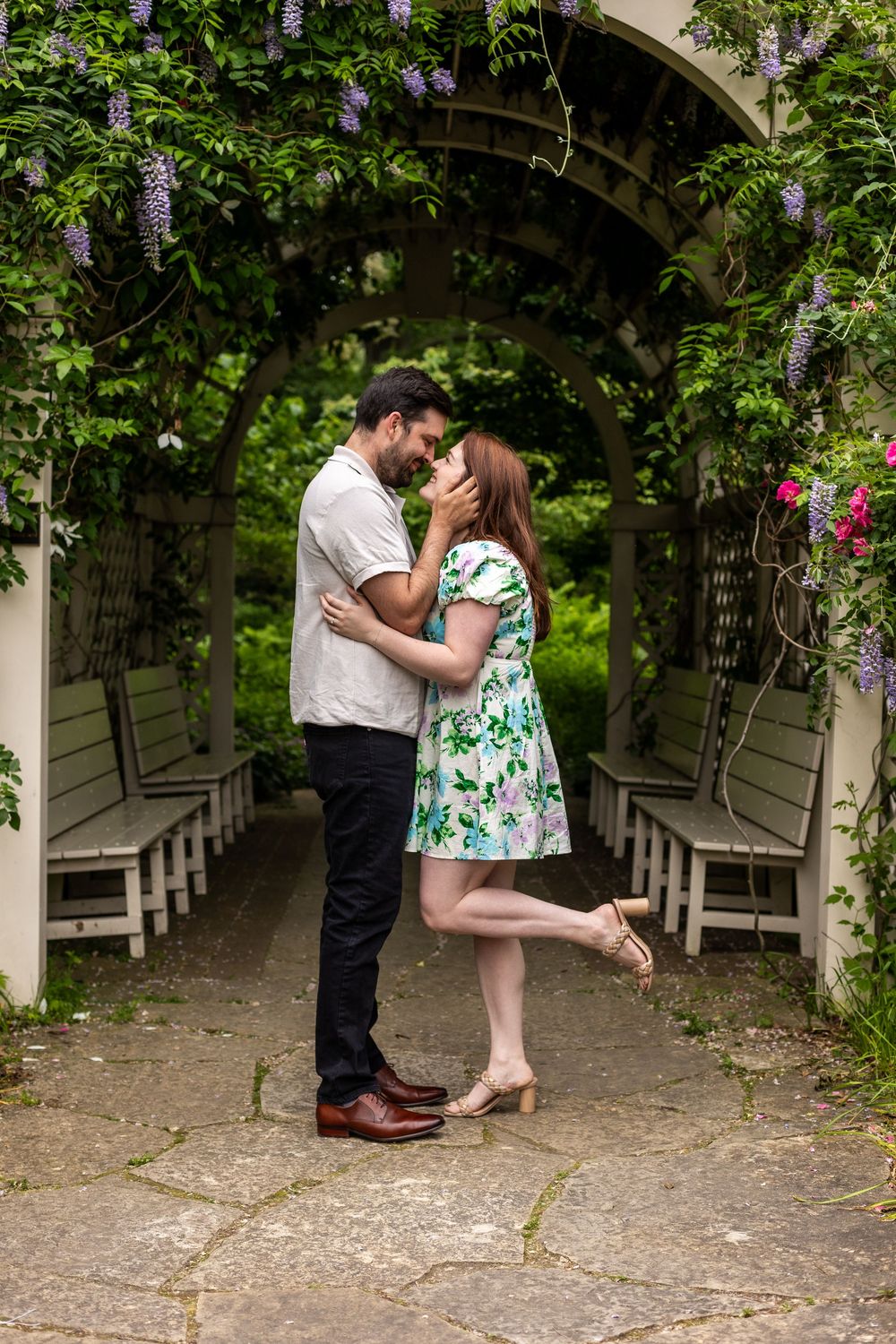 A romantic couple embraces in a lush garden archway with flowering vines and white benches creating a magical setting.