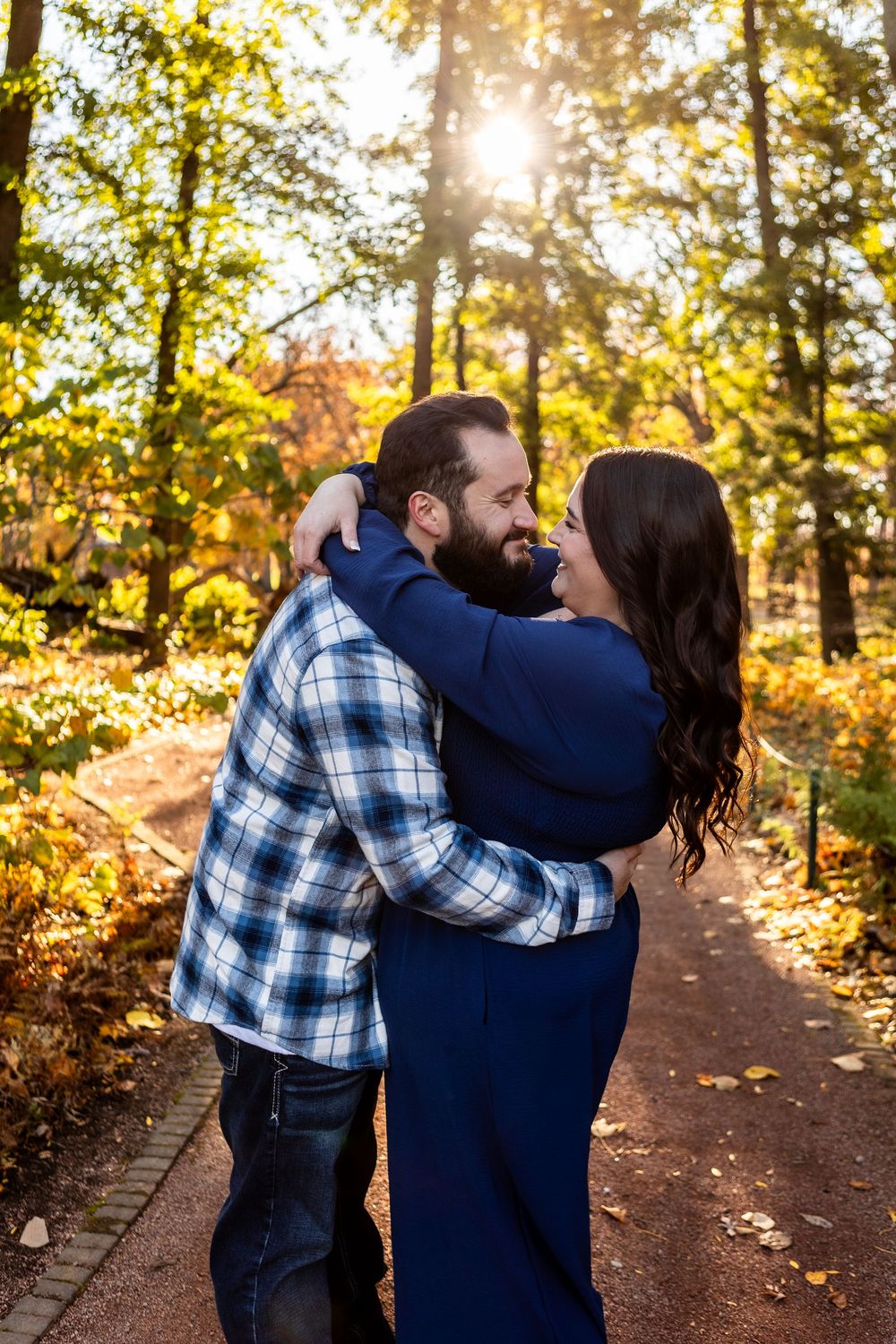 A couple embraces on an autumn path surrounded by golden leaves and sunlit trees in a romantic outdoor setting.