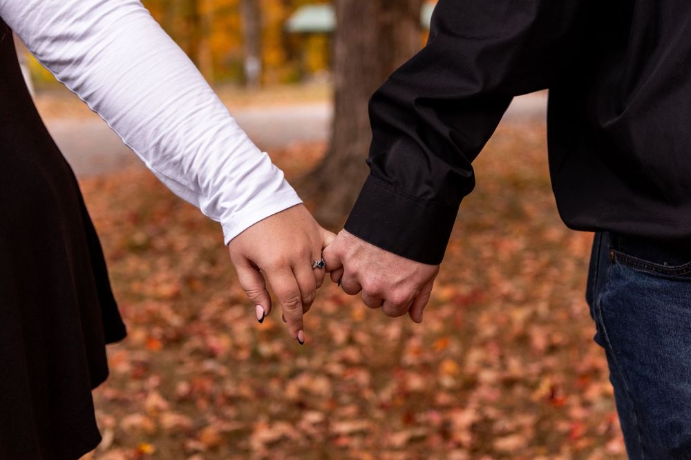 Two people hold hands while walking through fallen autumn leaves on a path during a fall day.