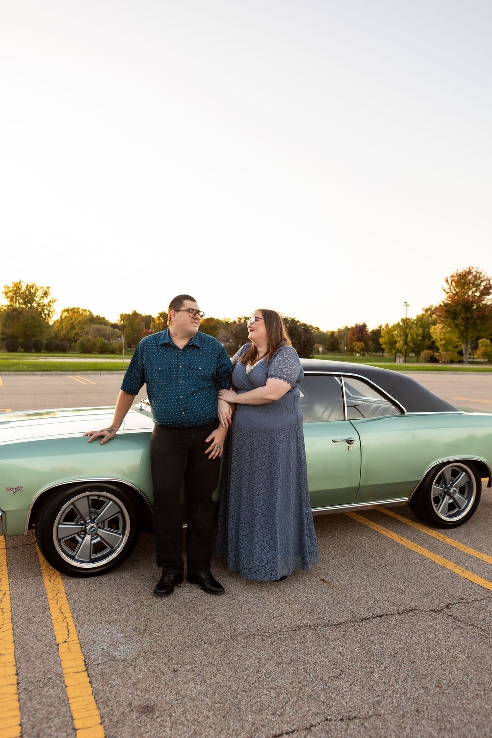 A couple poses with a classic mint green car in a parking lot during golden hour sunset.