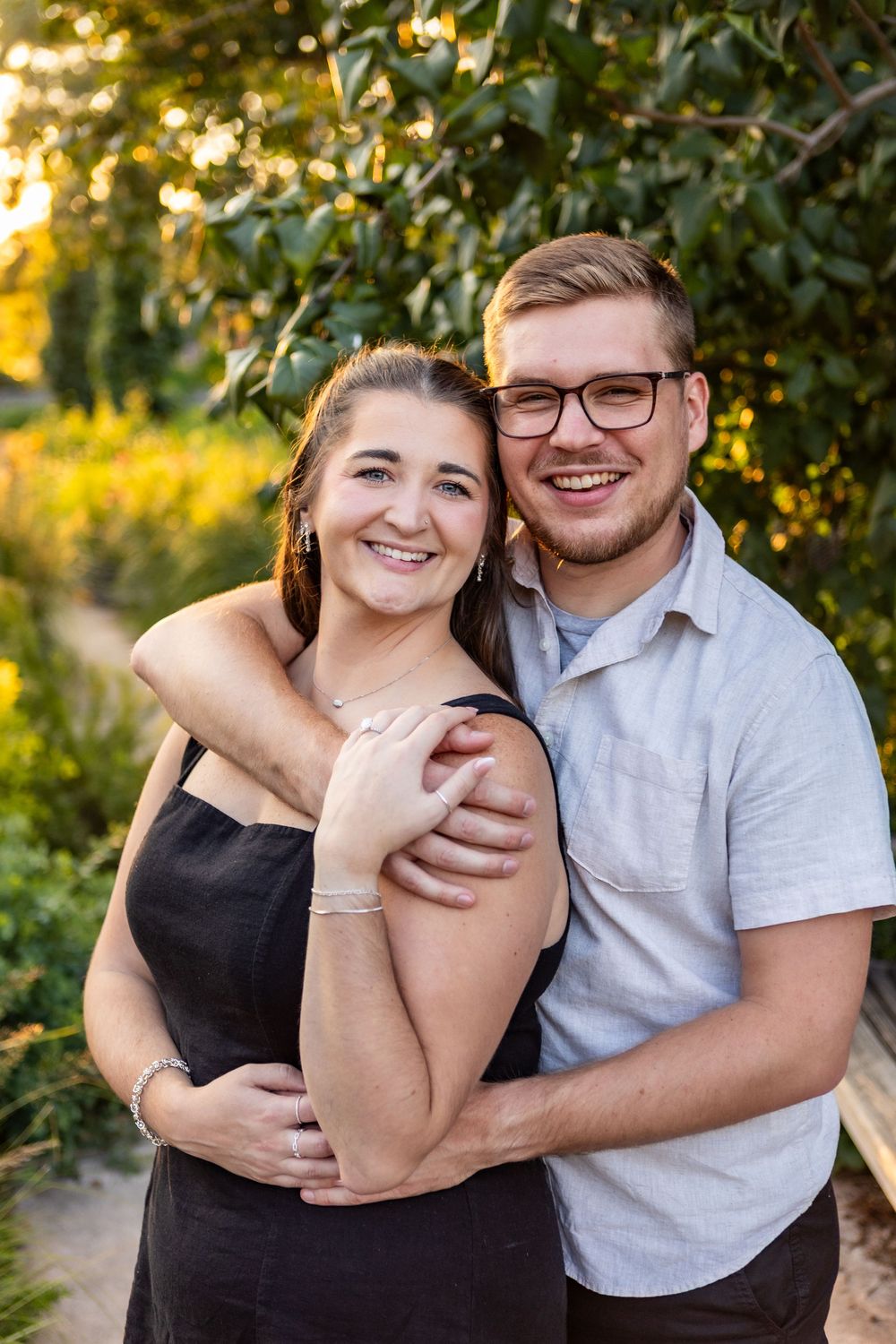 A couple embraces and laughs together during a sunset outdoor photo session with green foliage in the background.