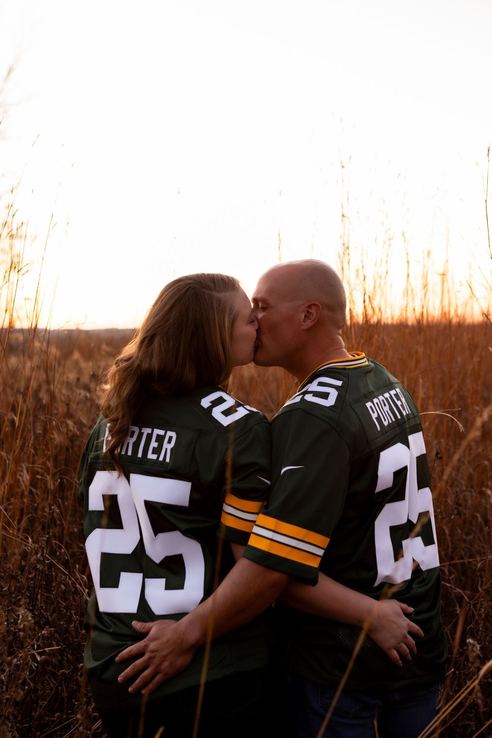 A couple wearing matching Green Bay Packers jerseys numbered 25 share a romantic sunset kiss in tall grass.