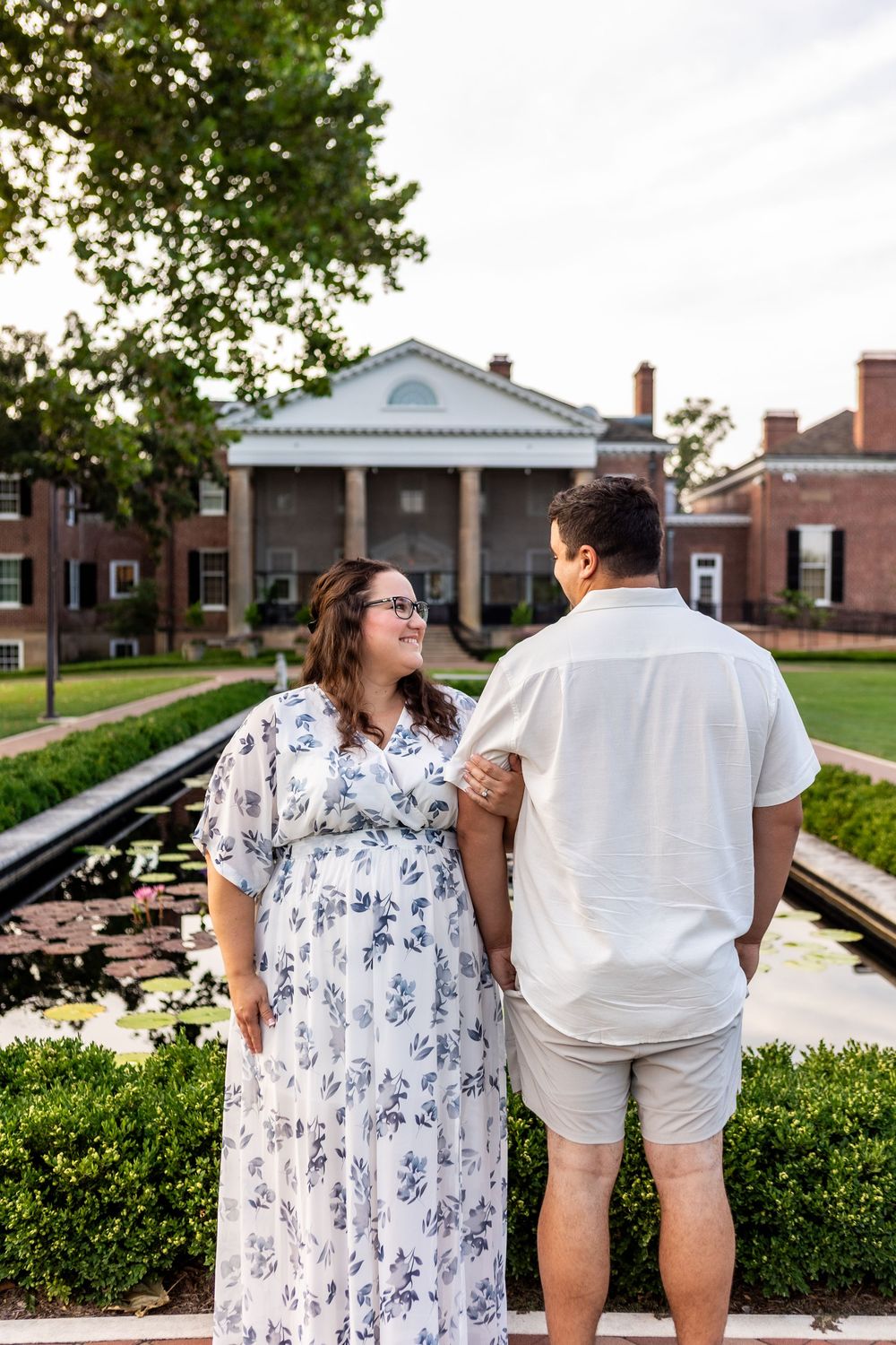 A couple in coordinating blue and white outfits pose together in a garden setting near brick buildings.