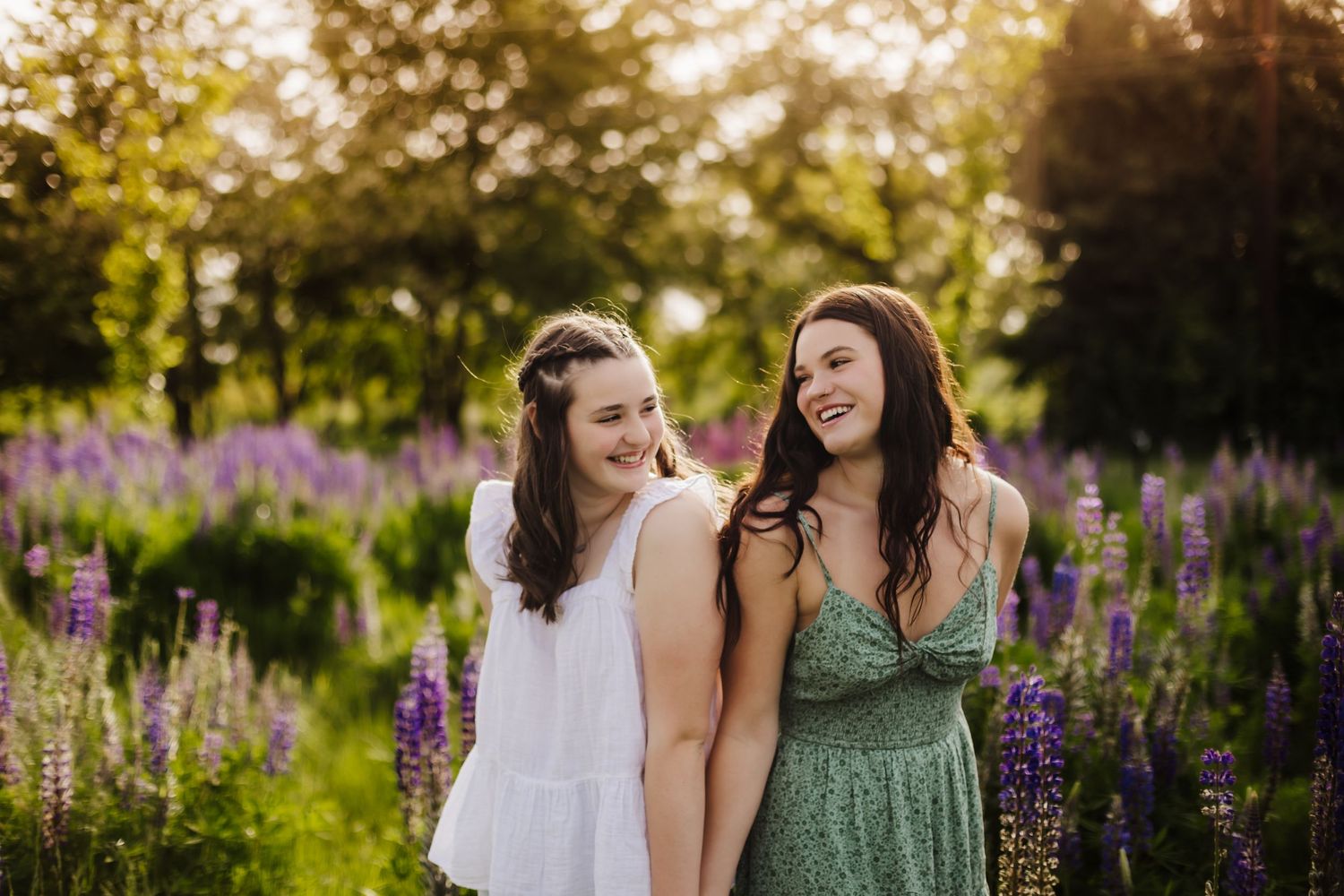 Two friends sharing laughter in a garden with blooming purple lupine flowers at sunset.