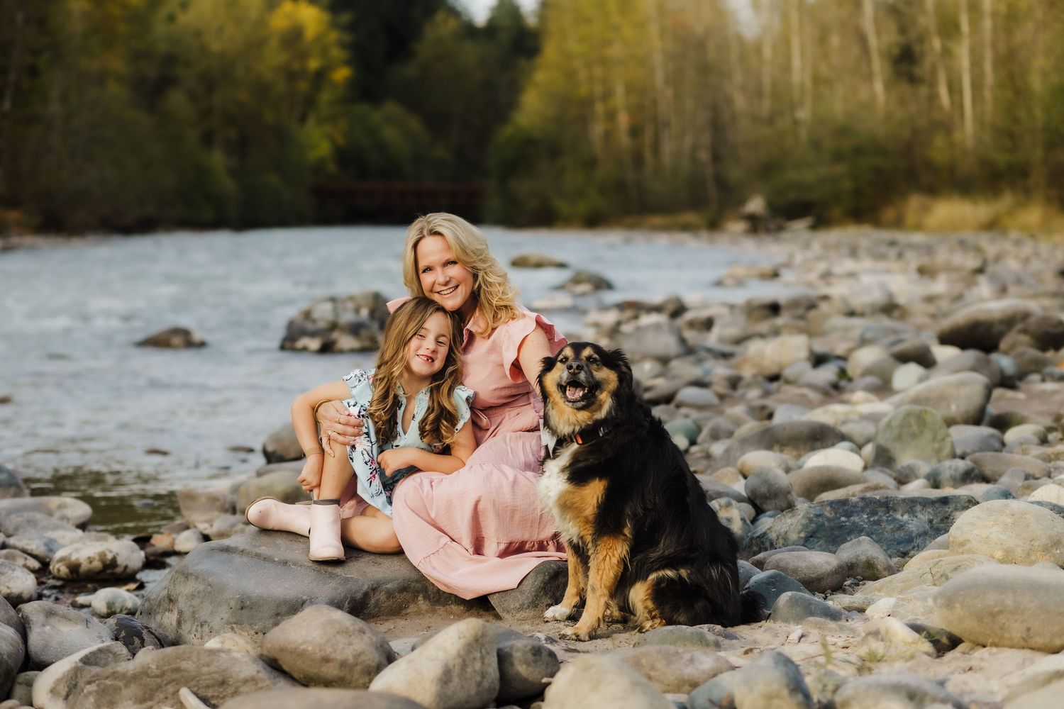 A couple sits on rocky riverbank with their German Shepherd dog during a scenic outdoor photo session.