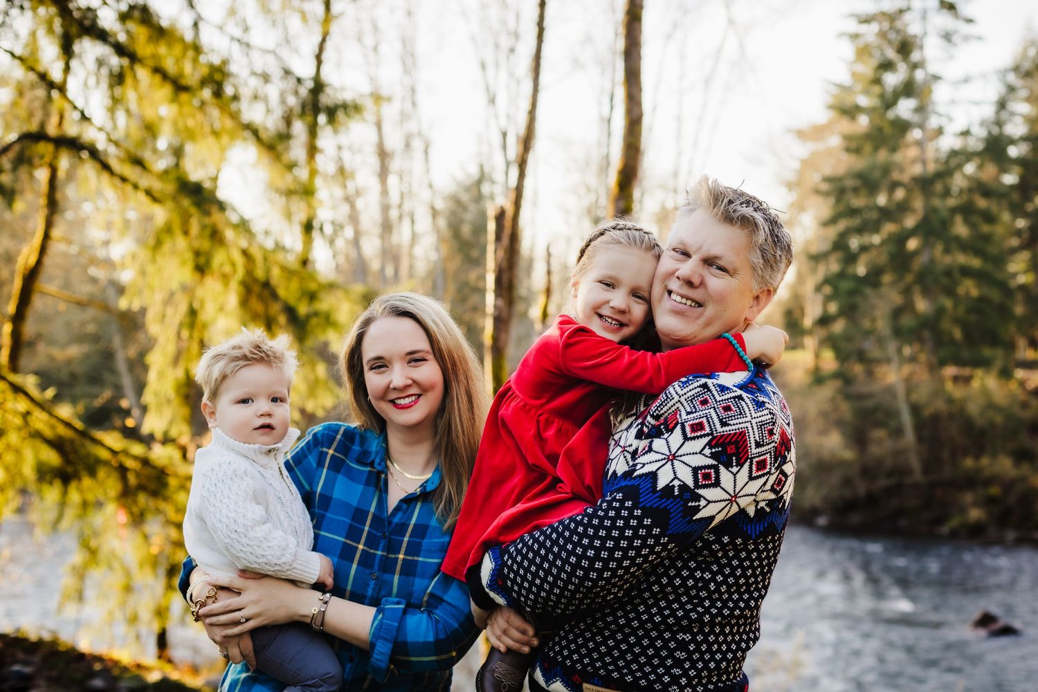 A happy family moment captured outdoors near a river with evergreen trees in the background.