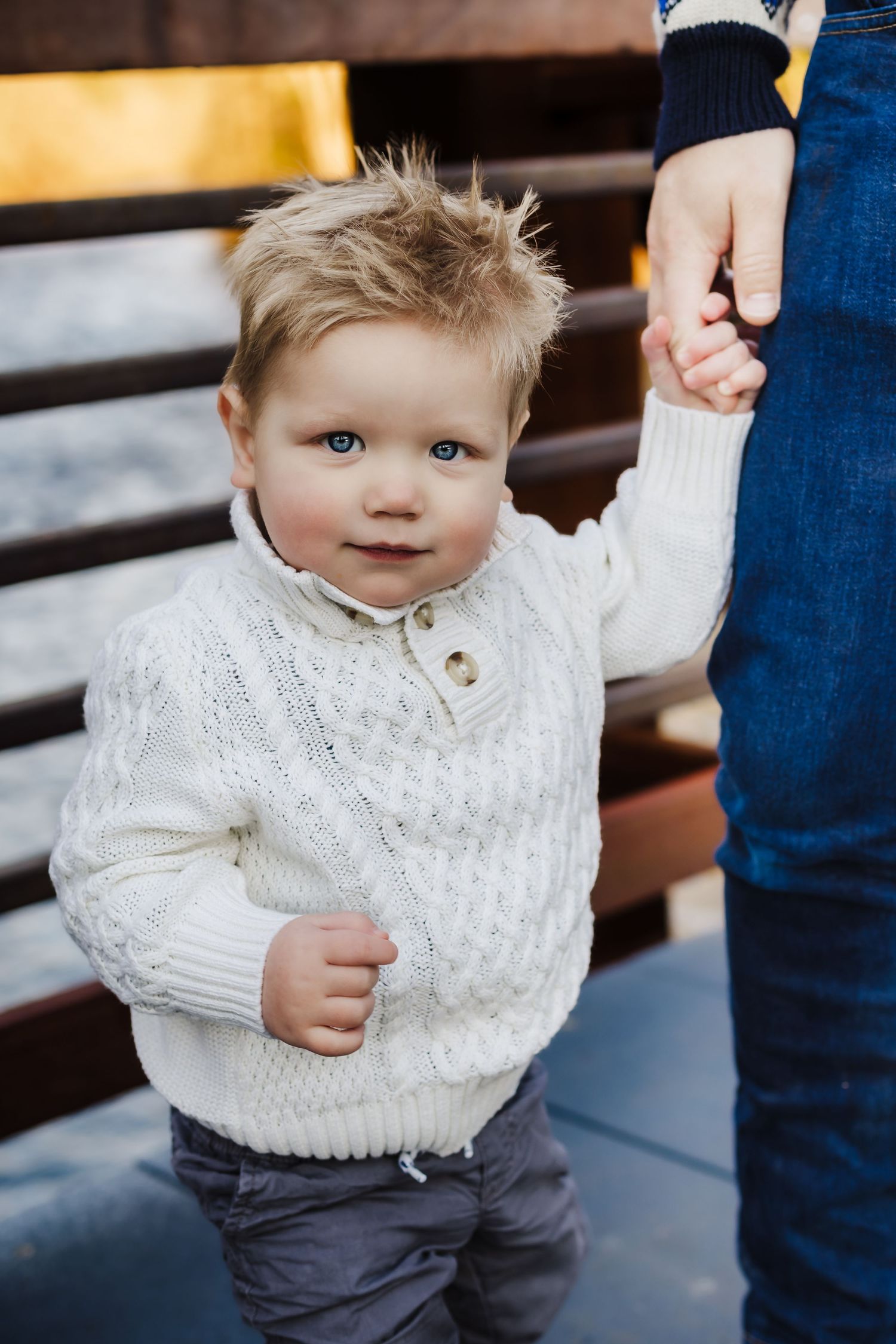 A toddler in a white cable knit sweater holding an adult's hand while standing on steps.