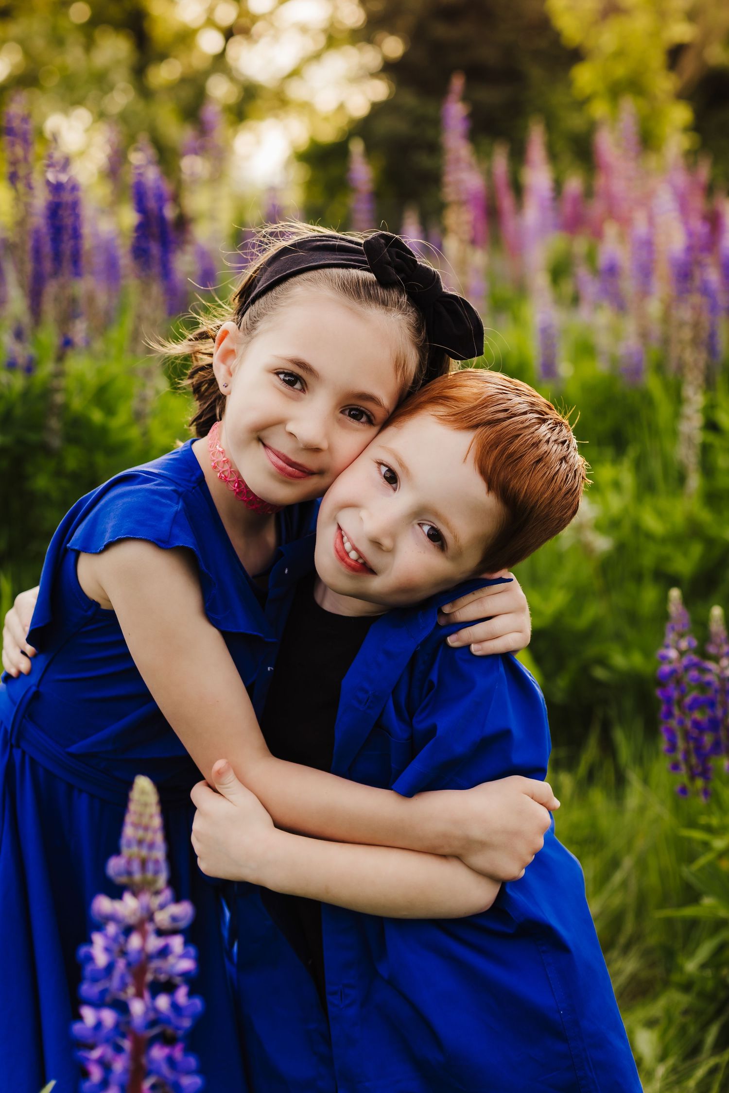 Two children in blue outfits embrace while smiling among purple lupine flowers.