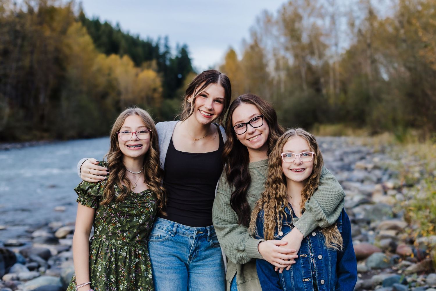 Four people pose together by a rocky riverbank with autumn trees in the background.