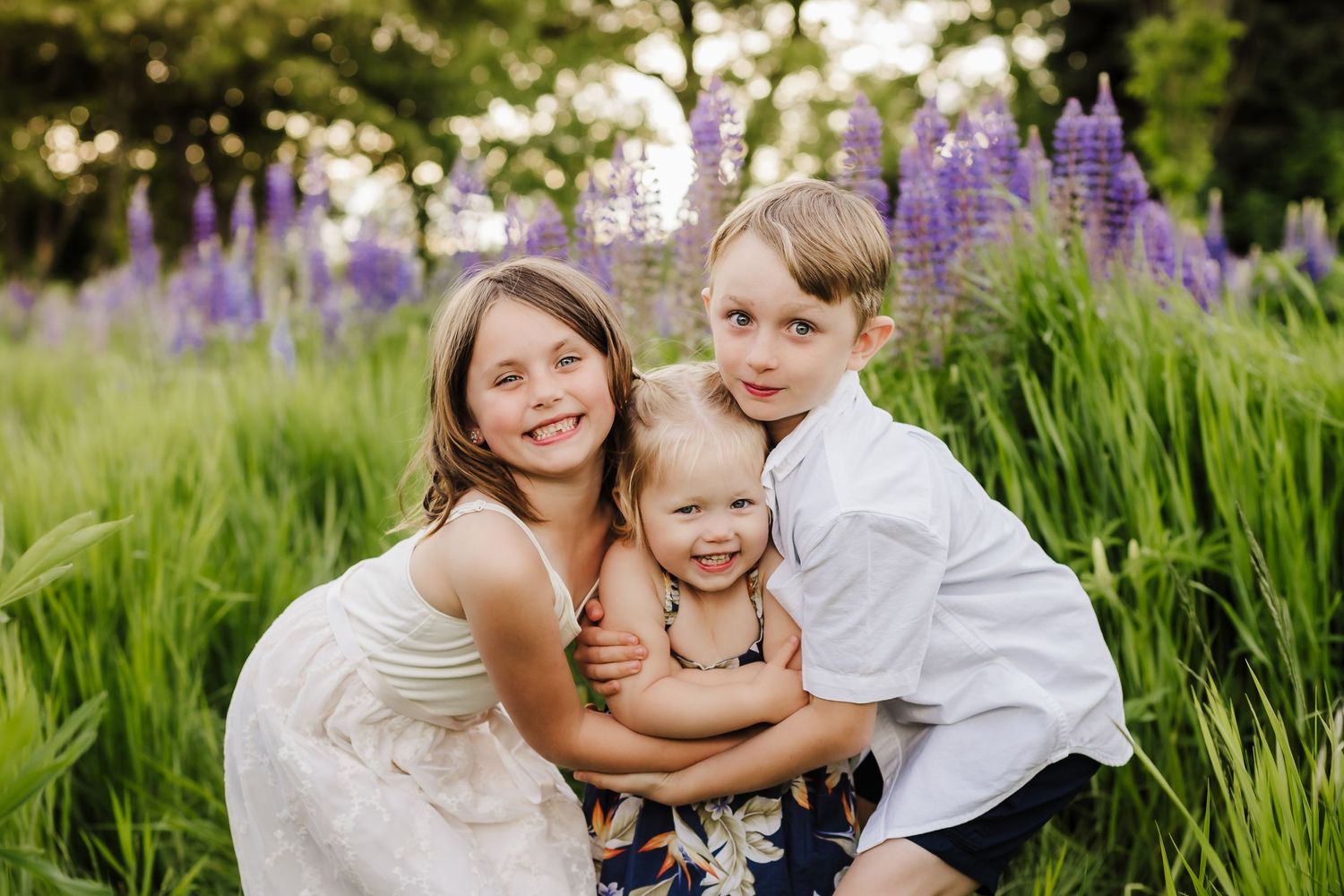 Three children share a group hug while posing in a field of purple lupine flowers.