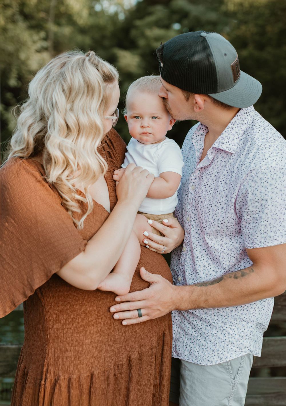 A couple in casual attire sharing an intimate moment with their baby outdoors in warm lighting.