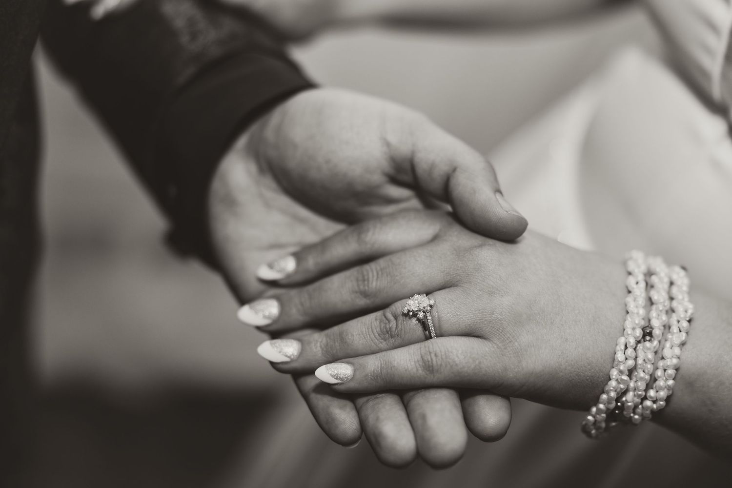 Black and white photo of hands clasped together in an intimate moment, with one hand wearing pearl bracelets.