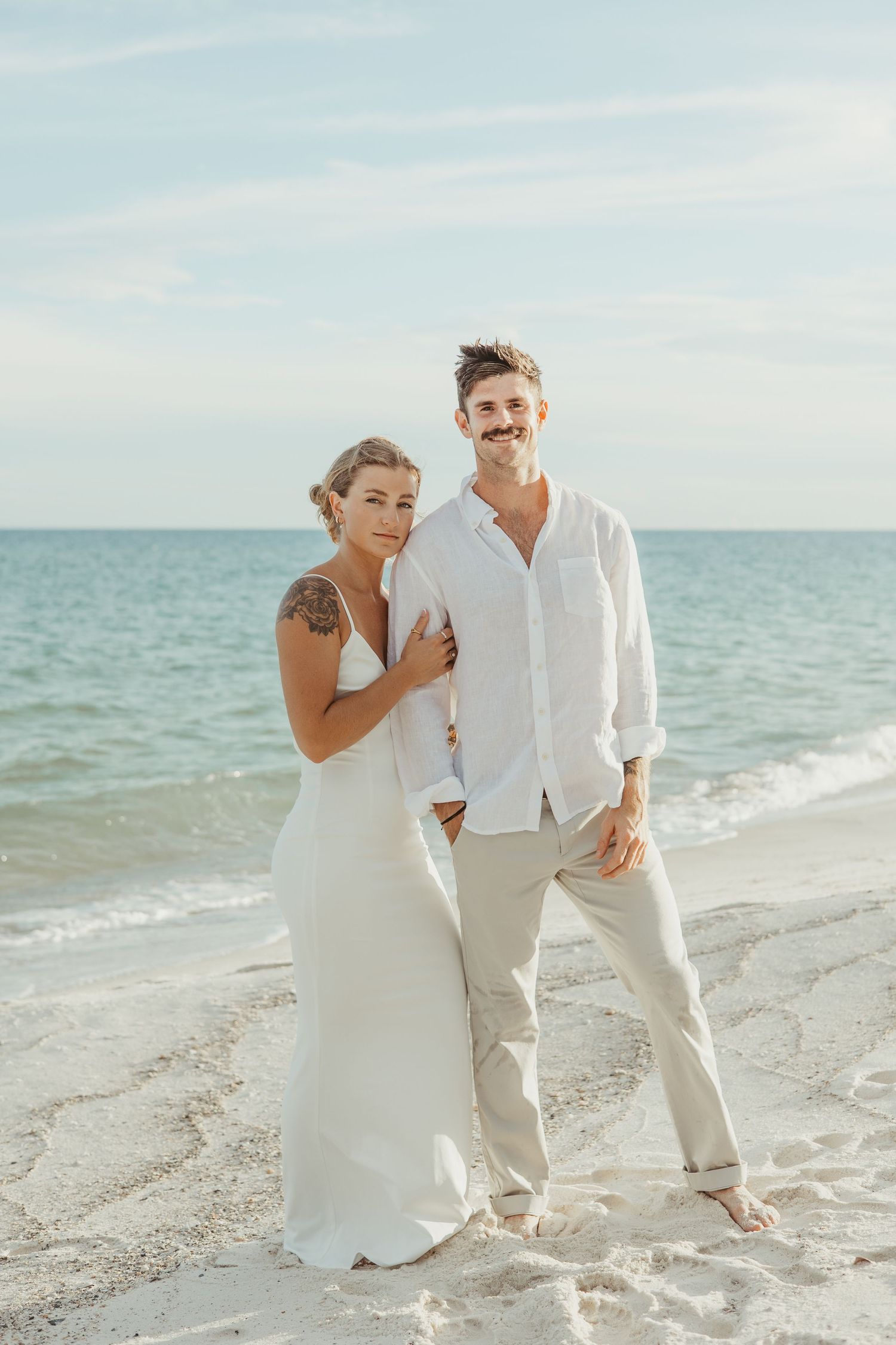 Newlywed couple laughing and walking together on a sunny beach in white wedding attire.