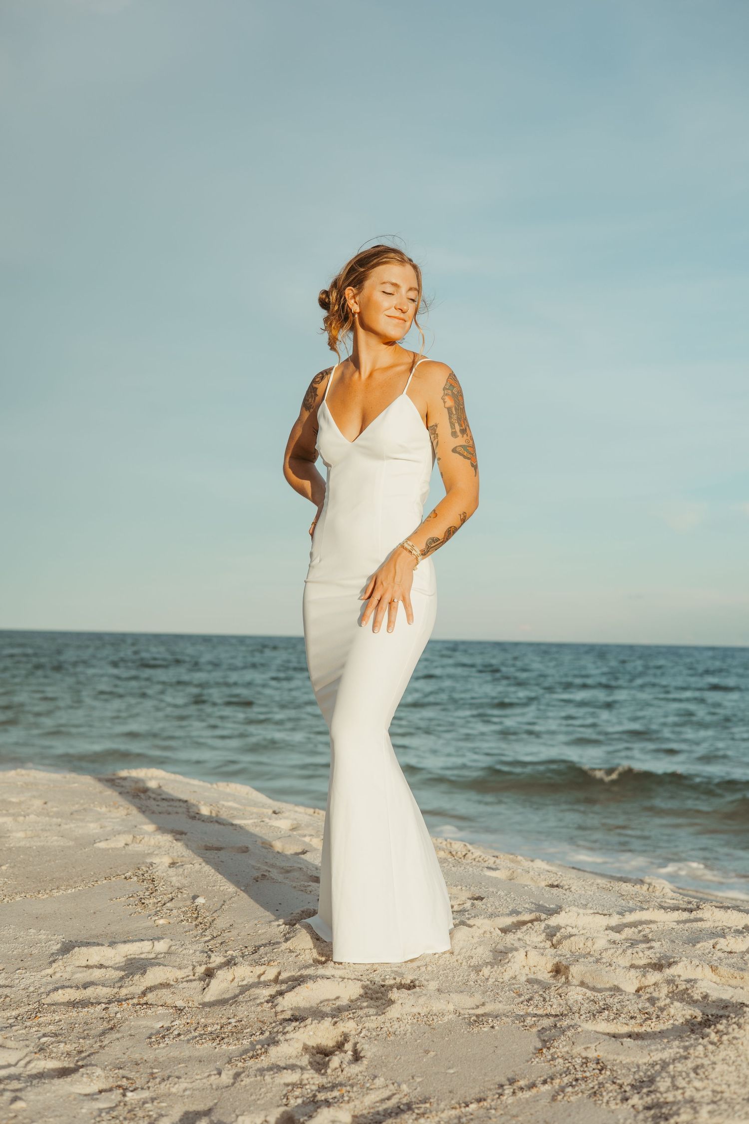 Woman in flowing white dress poses gracefully on sandy beach at sunset with ocean waves in background.