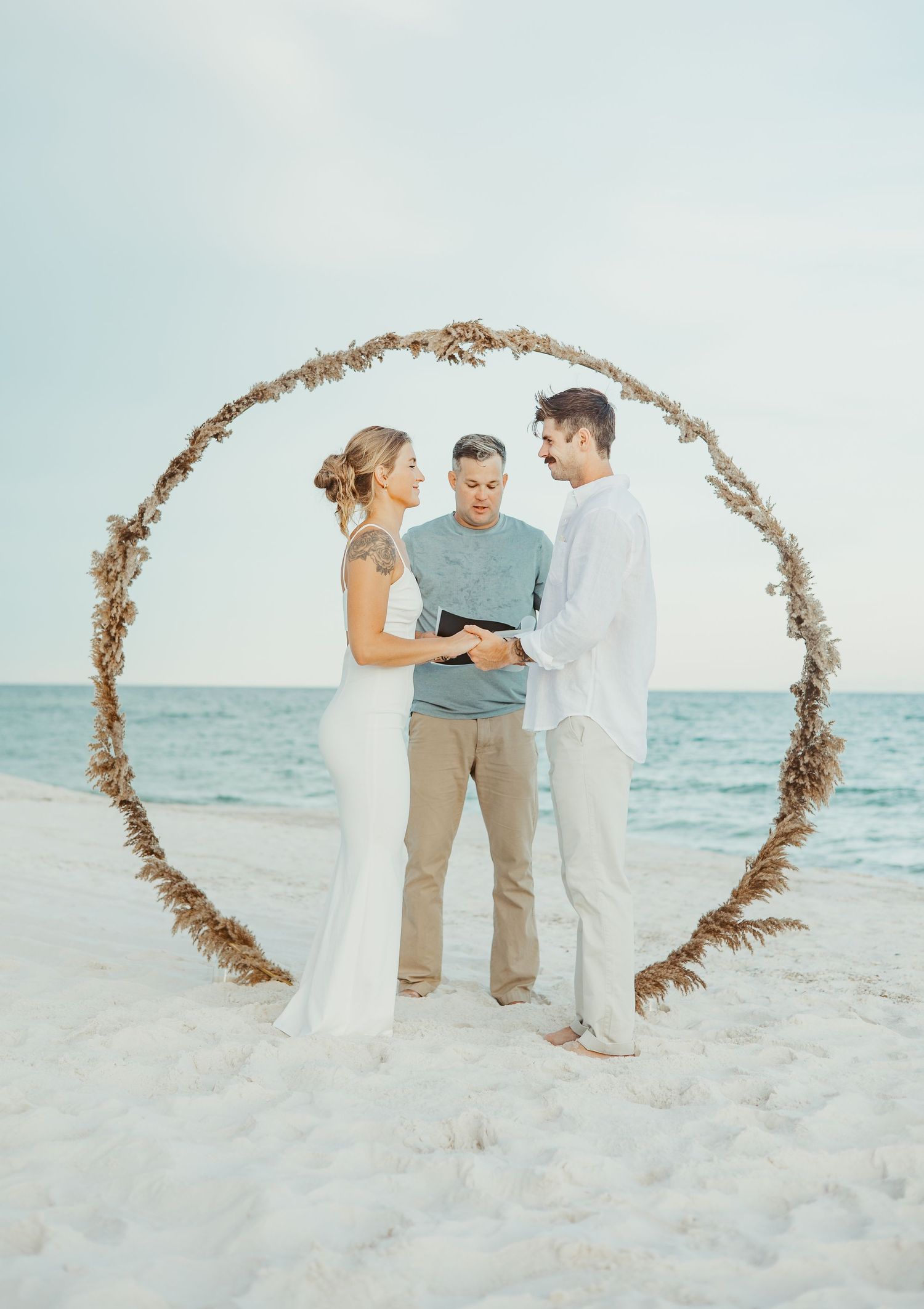 Beach wedding ceremony on white sand with rustic driftwood arch and ocean backdrop at sunset.