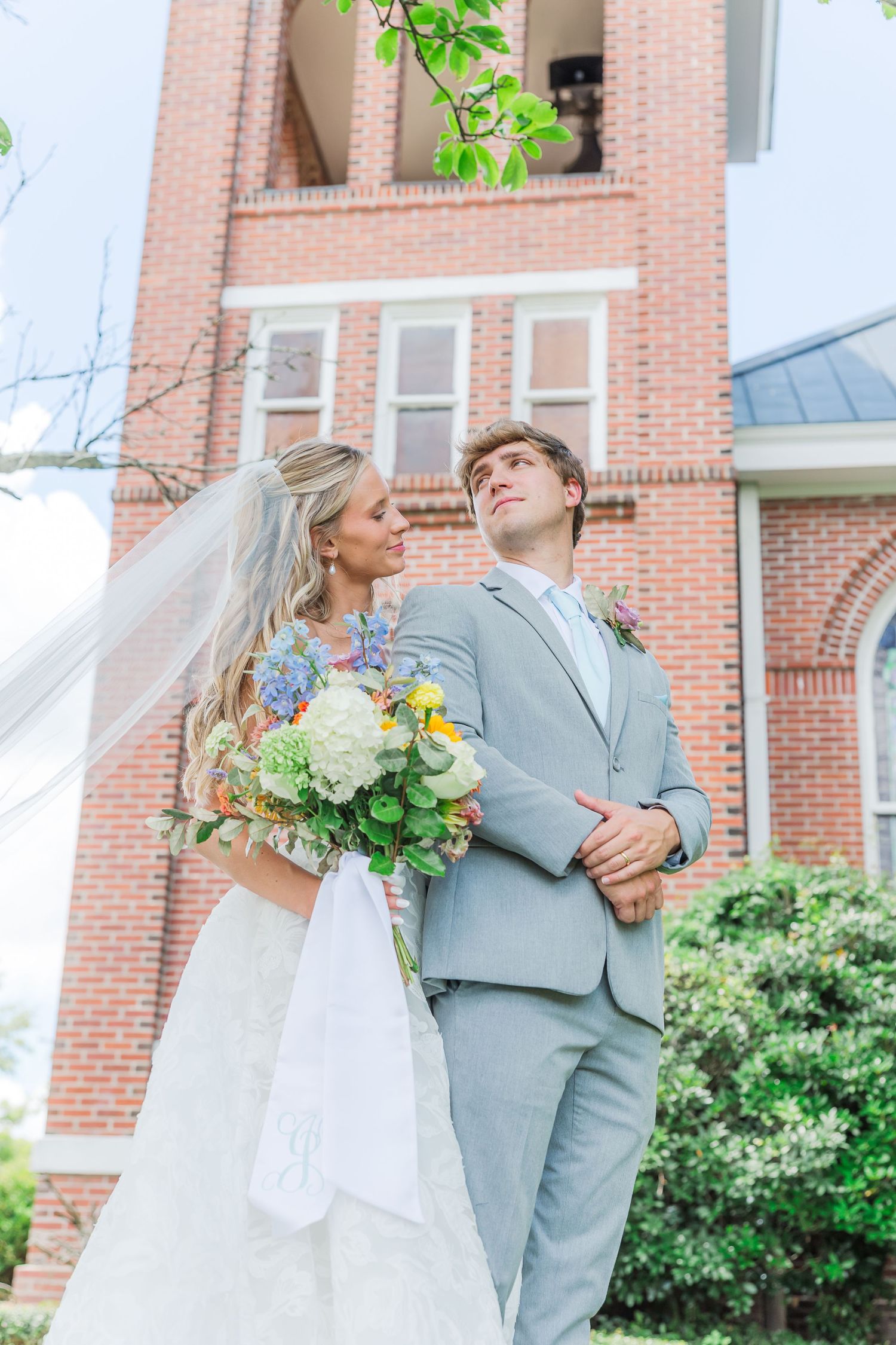 A couple in wedding attire poses playfully in front of a brick church.
