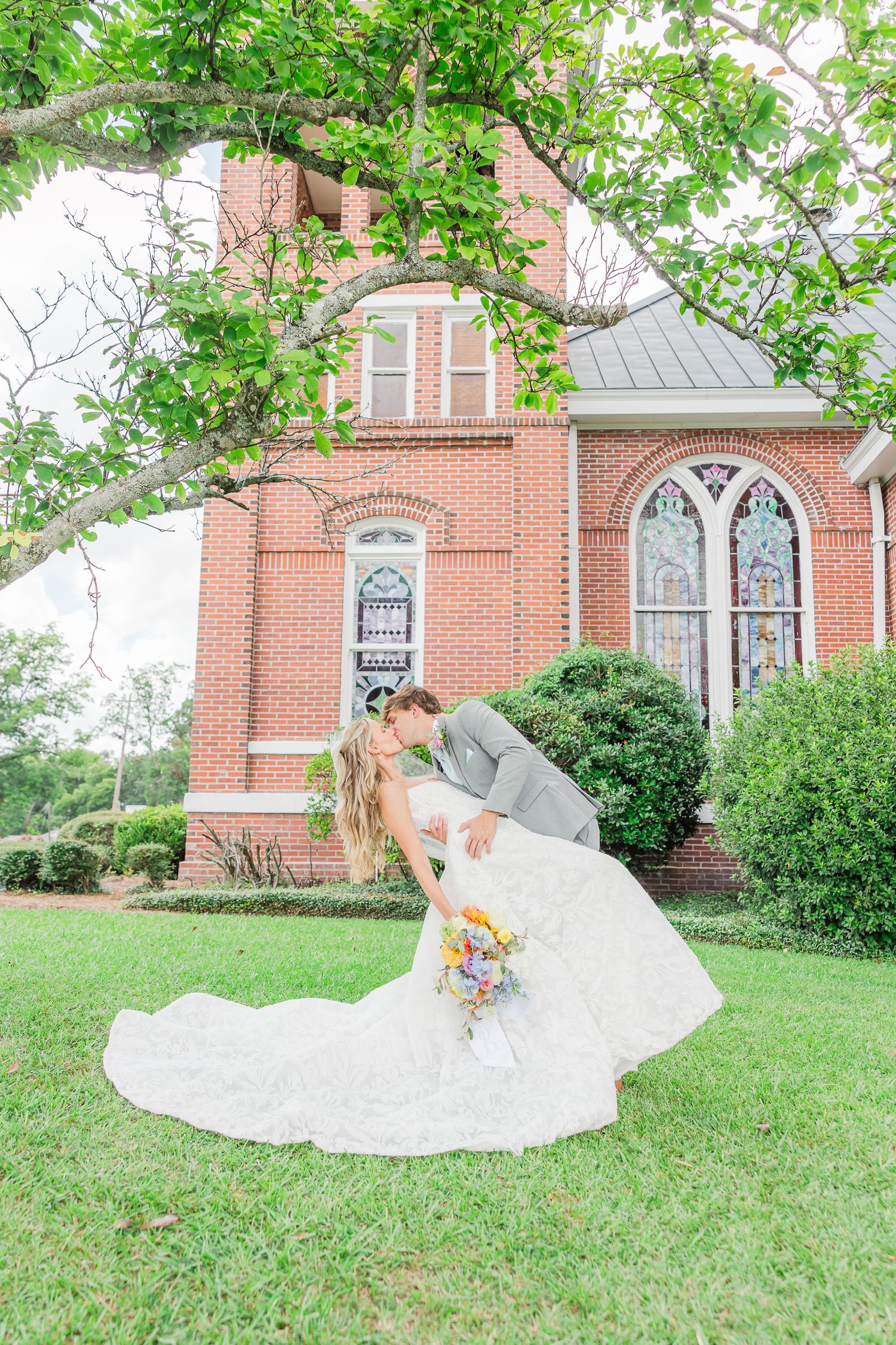 A flowing white wedding dress spreads across green grass with a brick church backdrop.