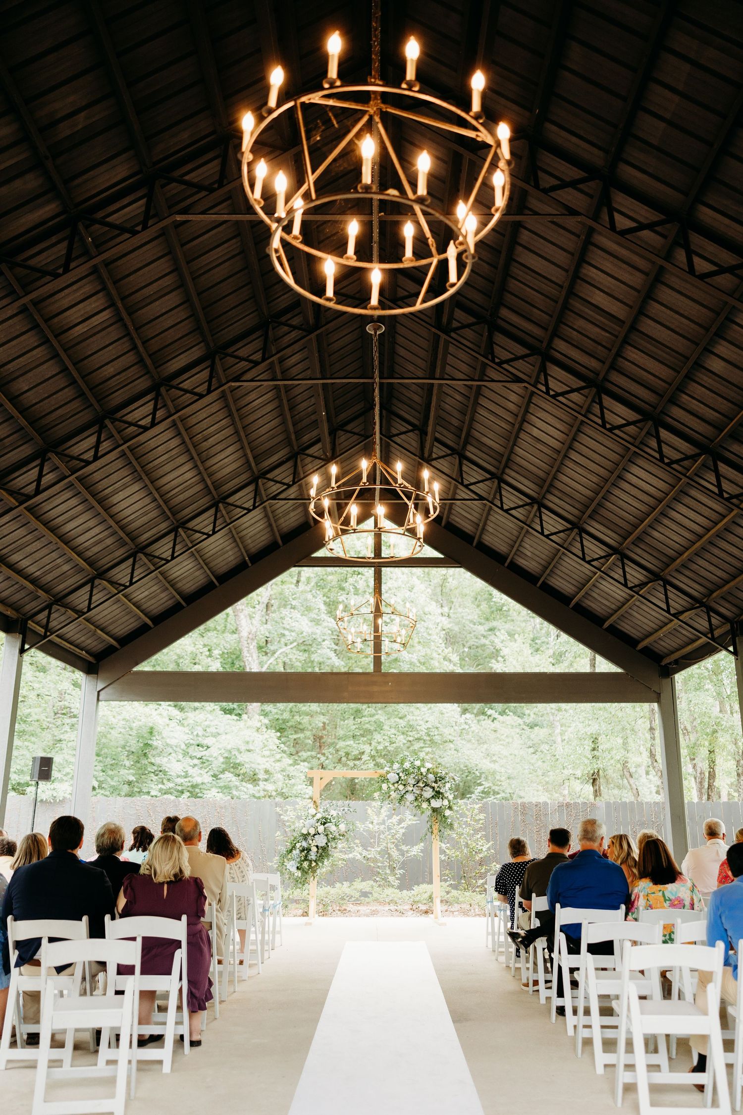 Rustic wooden pavilion with circular chandeliers and white chairs set up for an outdoor wedding ceremony.