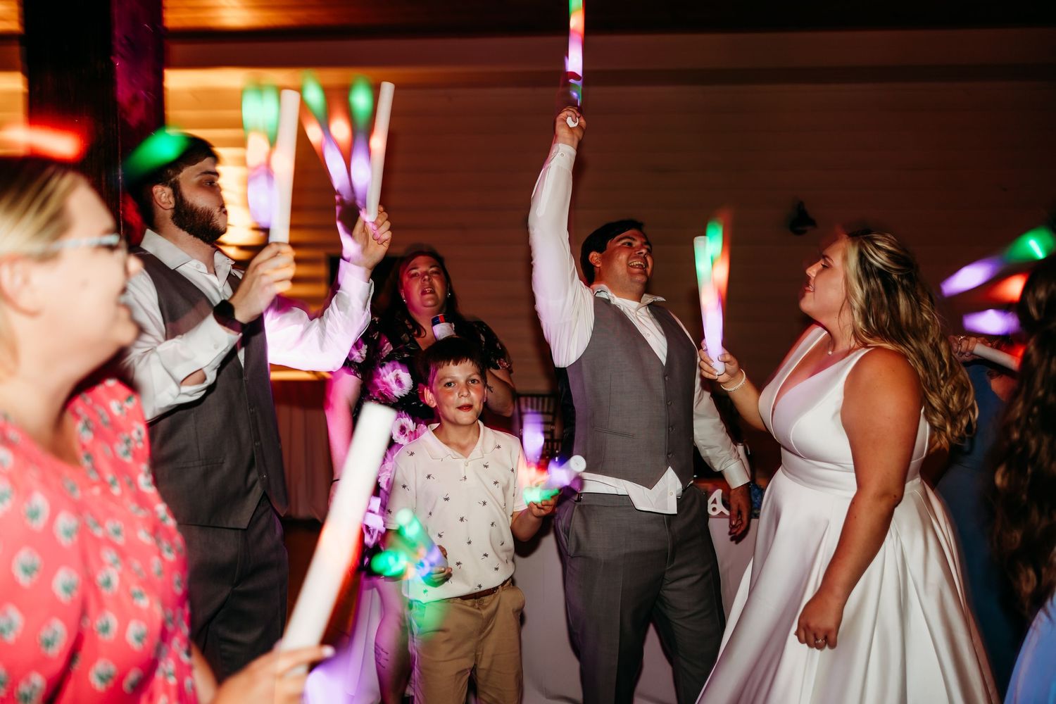 Wedding guests dance and celebrate on a dimly lit dance floor with colorful party lights in the background.
