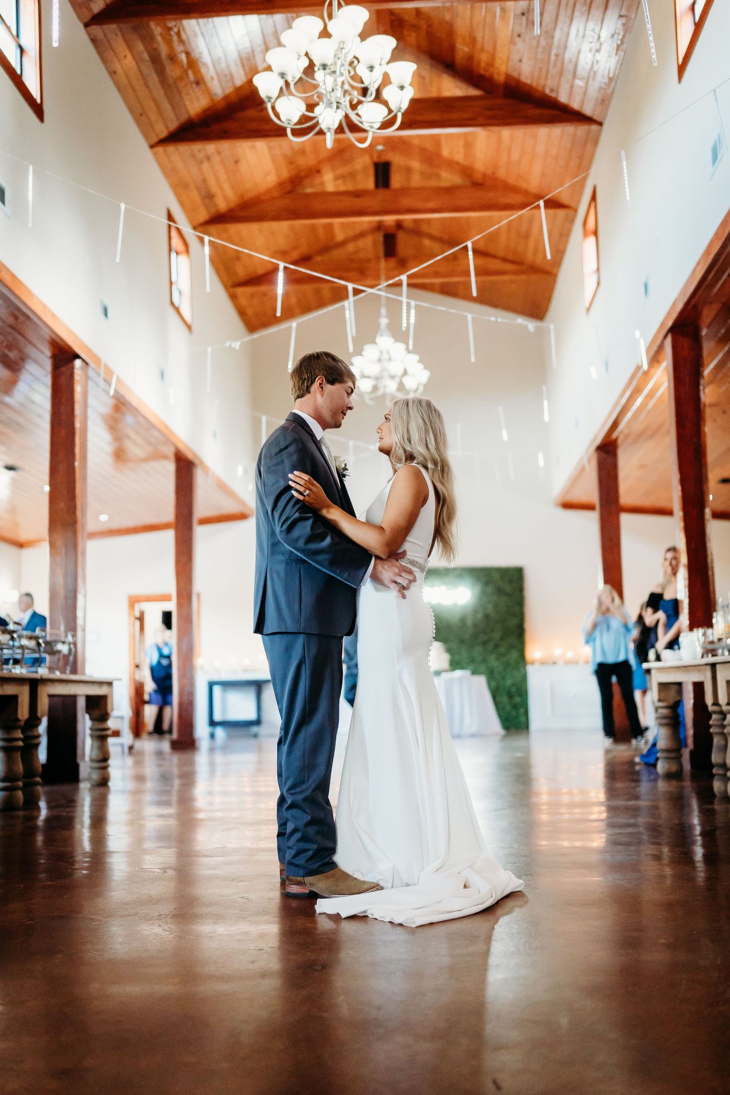 A couple shares a romantic dance in a rustic venue with wooden beams and chandeliers overhead.