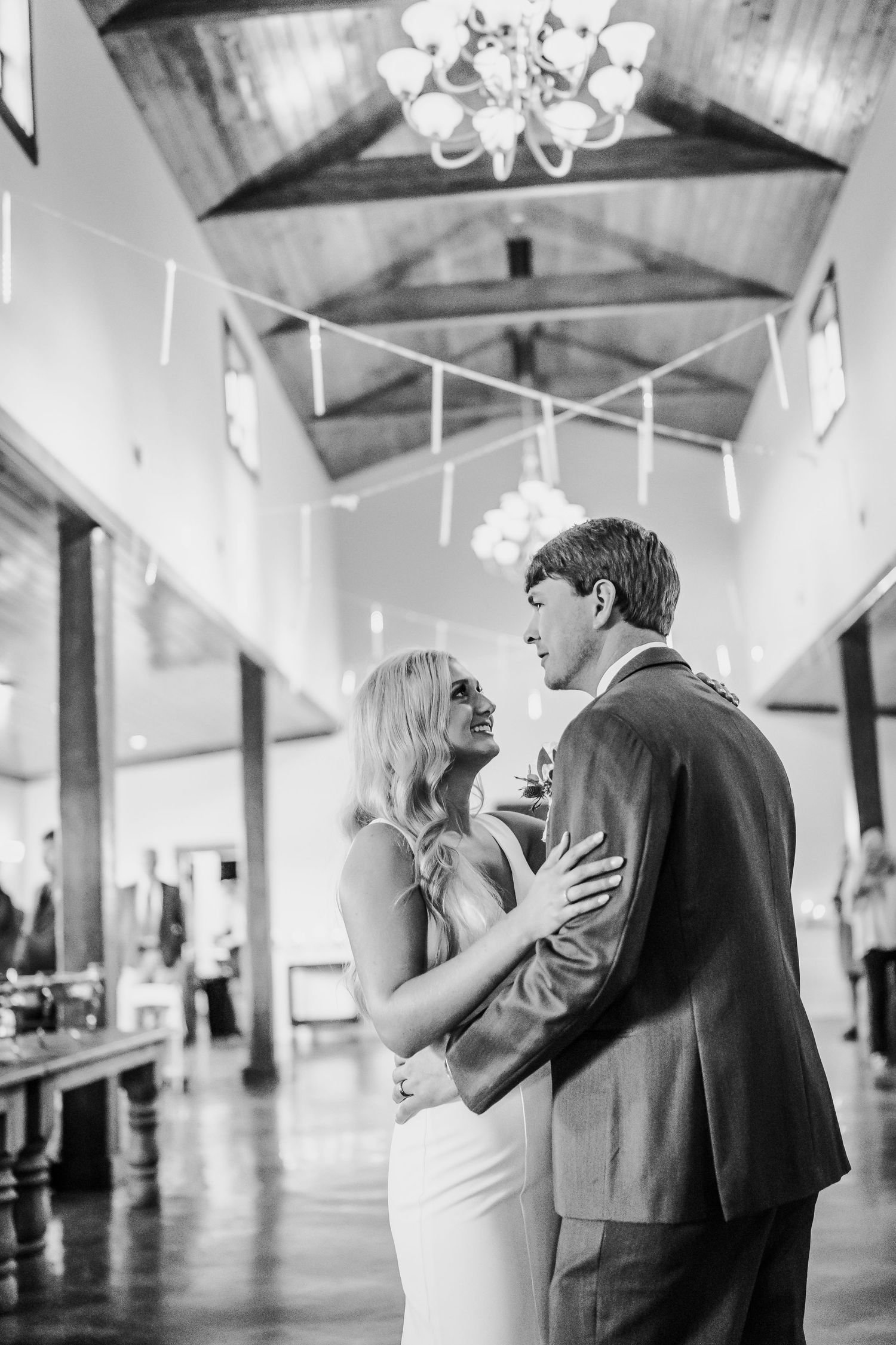 A couple shares an intimate dance moment in a rustic venue with exposed wooden beams and elegant chandeliers.