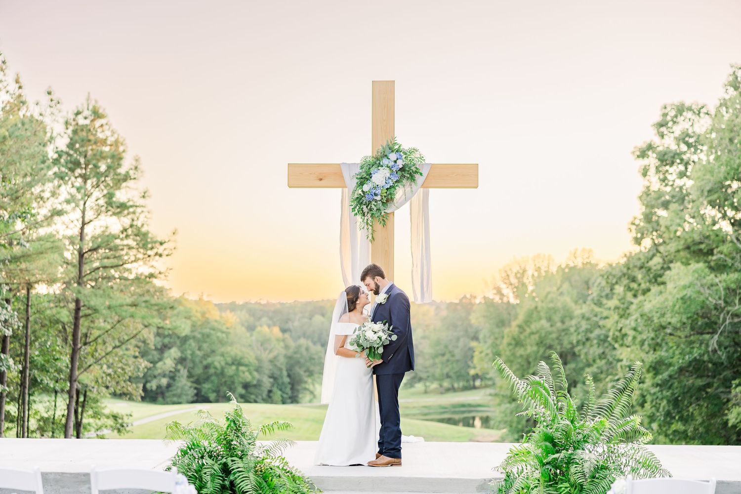 A romantic sunset wedding ceremony scene with a wooden cross and natural landscape backdrop.