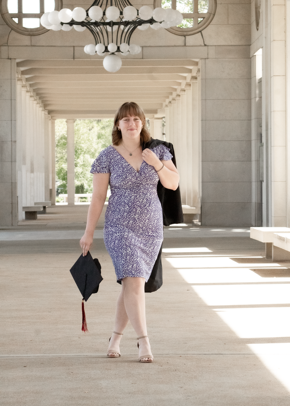 Person in floral dress and black jacket poses in sunlit colonnade with white pillars and architectural details.