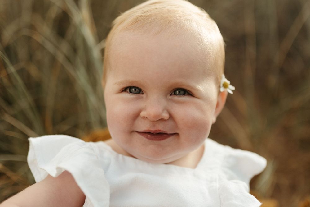 A happy baby wearing a white dress smiles brightly while sitting outdoors against a natural brown background.