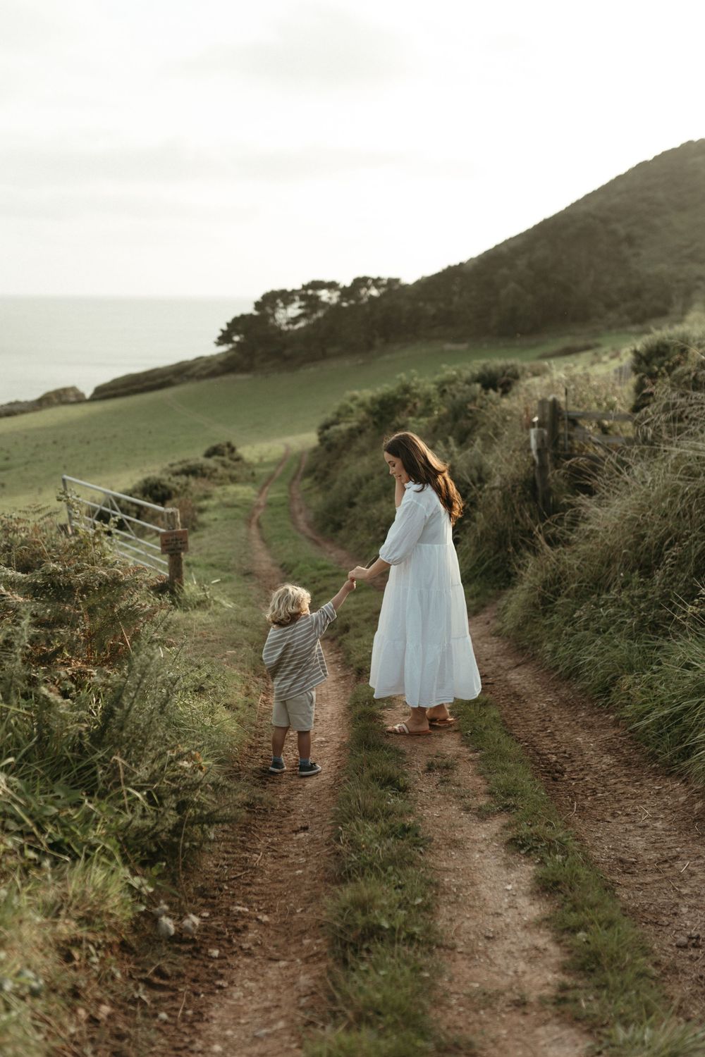 A child and figure in a white dress walk hand in hand along a coastal dirt path against rolling green hills and ocean.