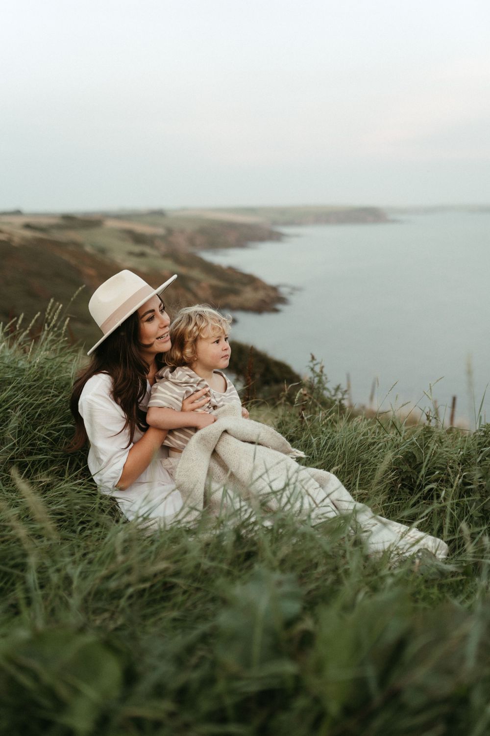 Two people relaxing on grassy coastal cliffs overlooking a serene ocean view while wearing white summer outfits.