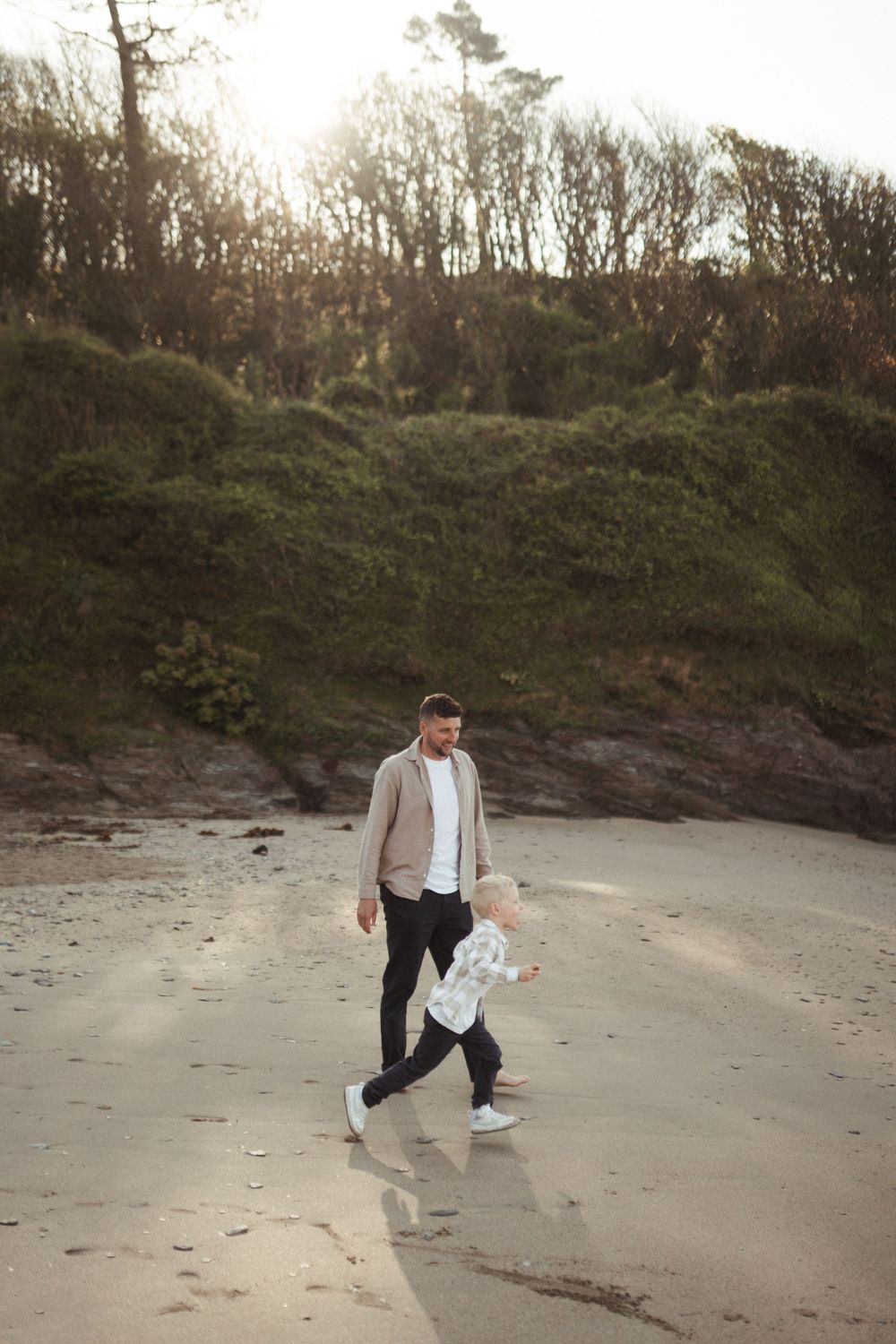 Someone walking along a sandy beach with trees and cliffs in the background at sunset while holding a water bottle.