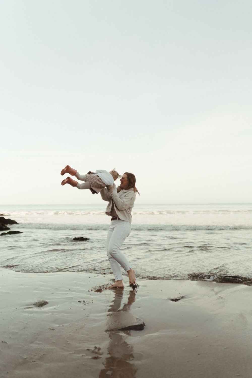 Two people in casual white clothes playing and spinning on a misty beach at sunset with waves crashing in the background.