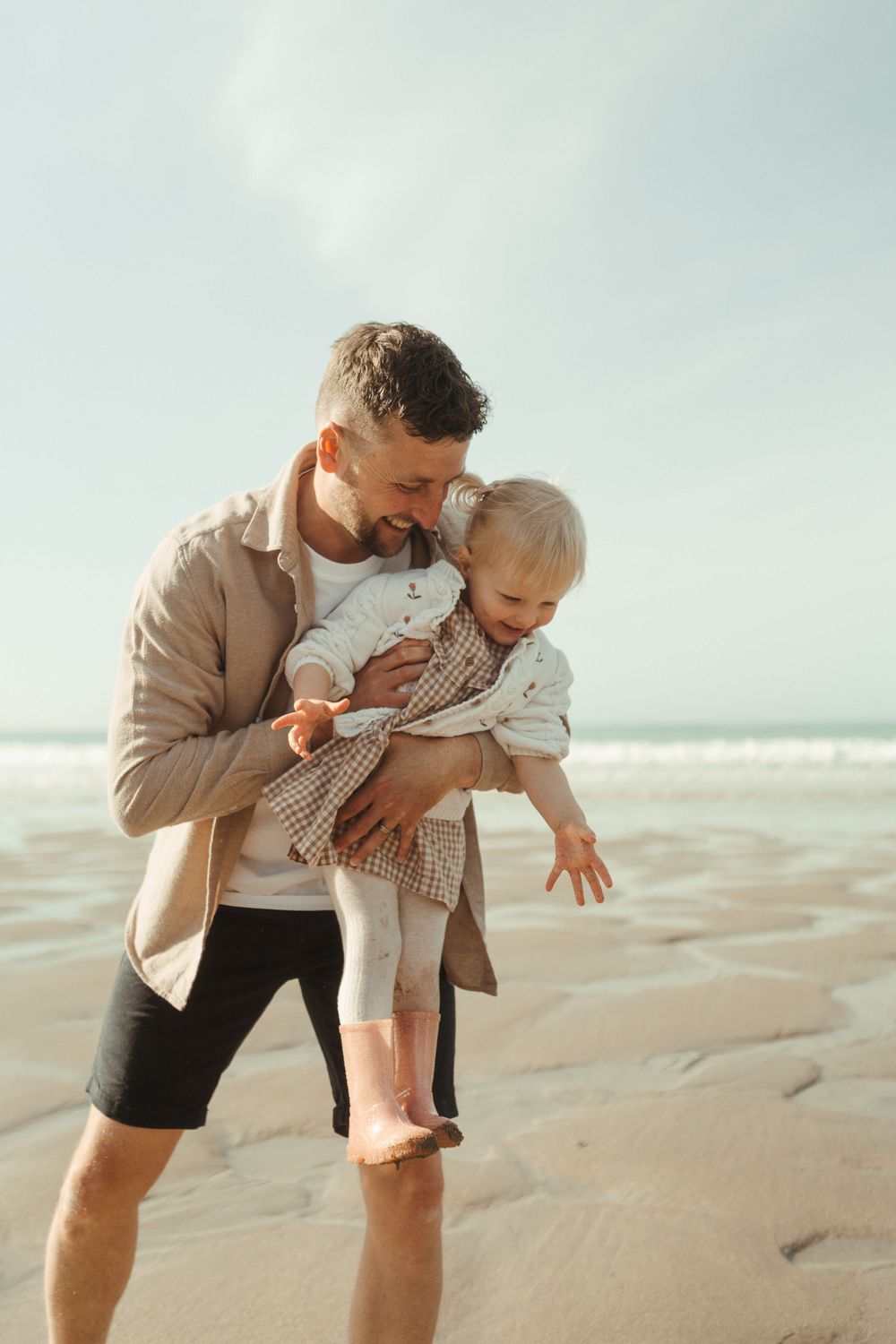 A parent and toddler share a playful moment together on a sunny beach with ocean waves in the background.