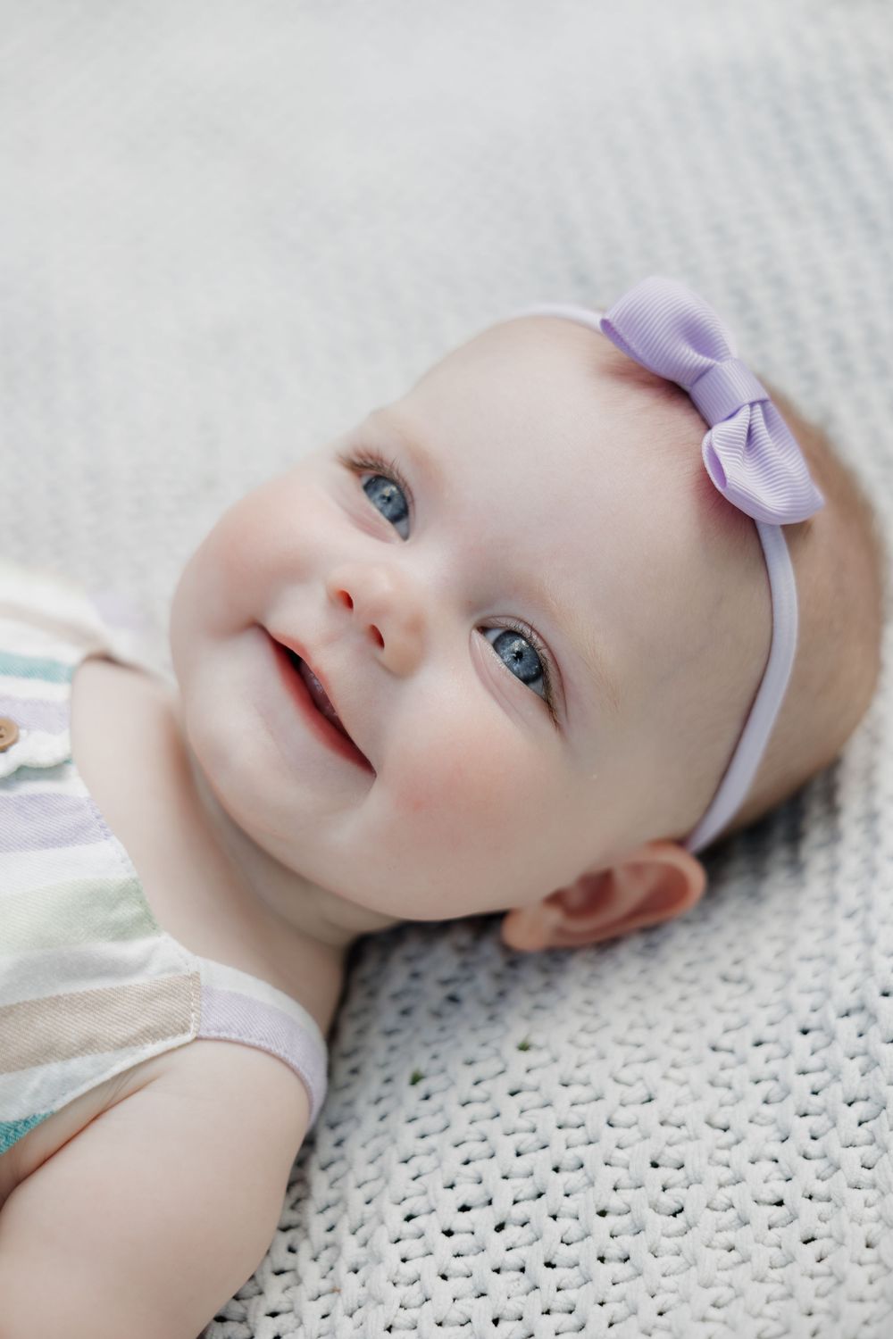 Smiling baby girl with blue eyes wearing purple bow headband lying on white knit blanket.