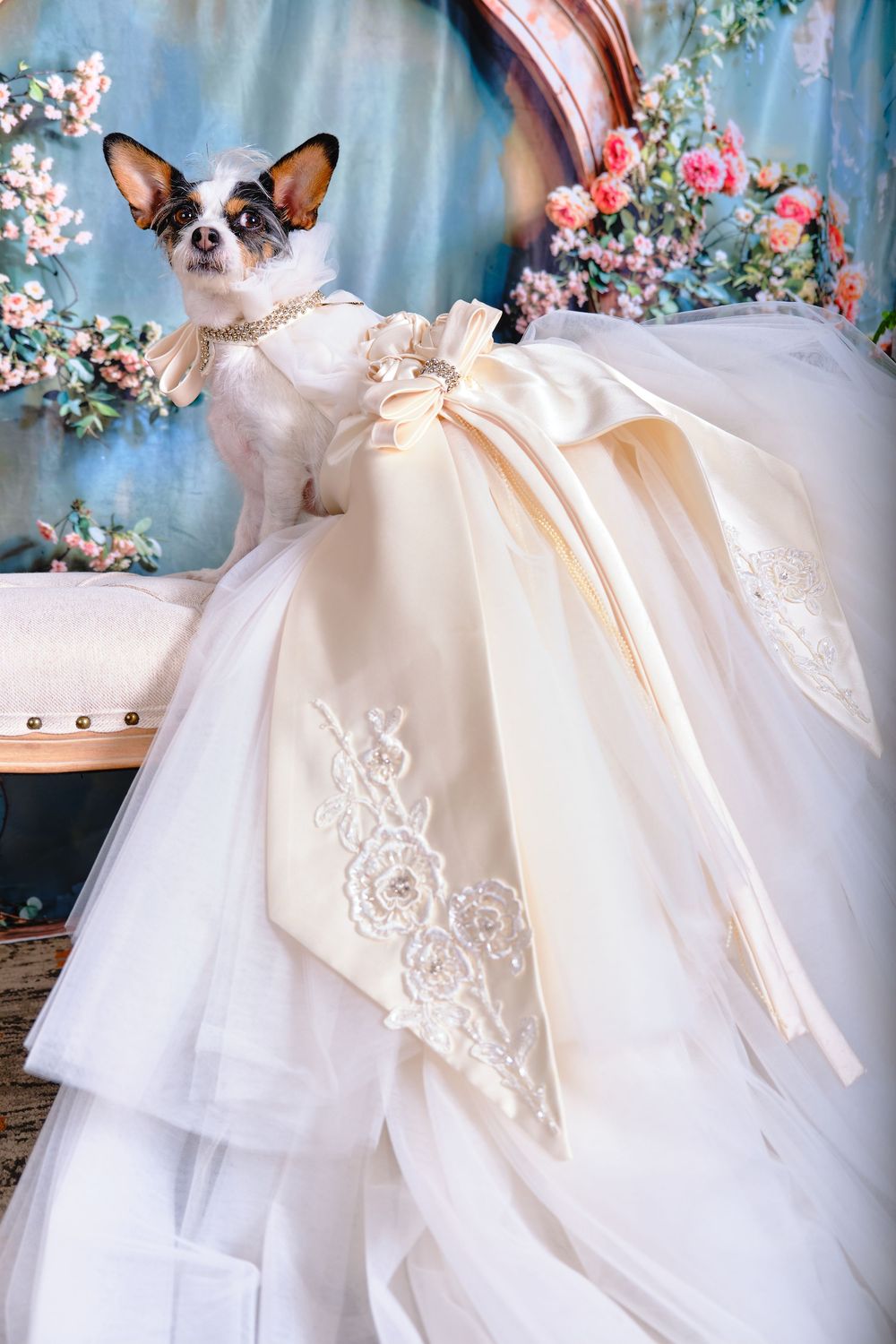 Elegant white tulle and lace wedding dress displayed with trailing bow details in floral backdrop setting.
