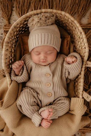 Sleeping baby in beige knitted outfit and matching hat resting peacefully in woven basket.