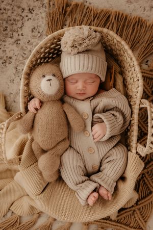 Baby in knitted beige outfit sleeping in basket with stuffed teddy bear companion.