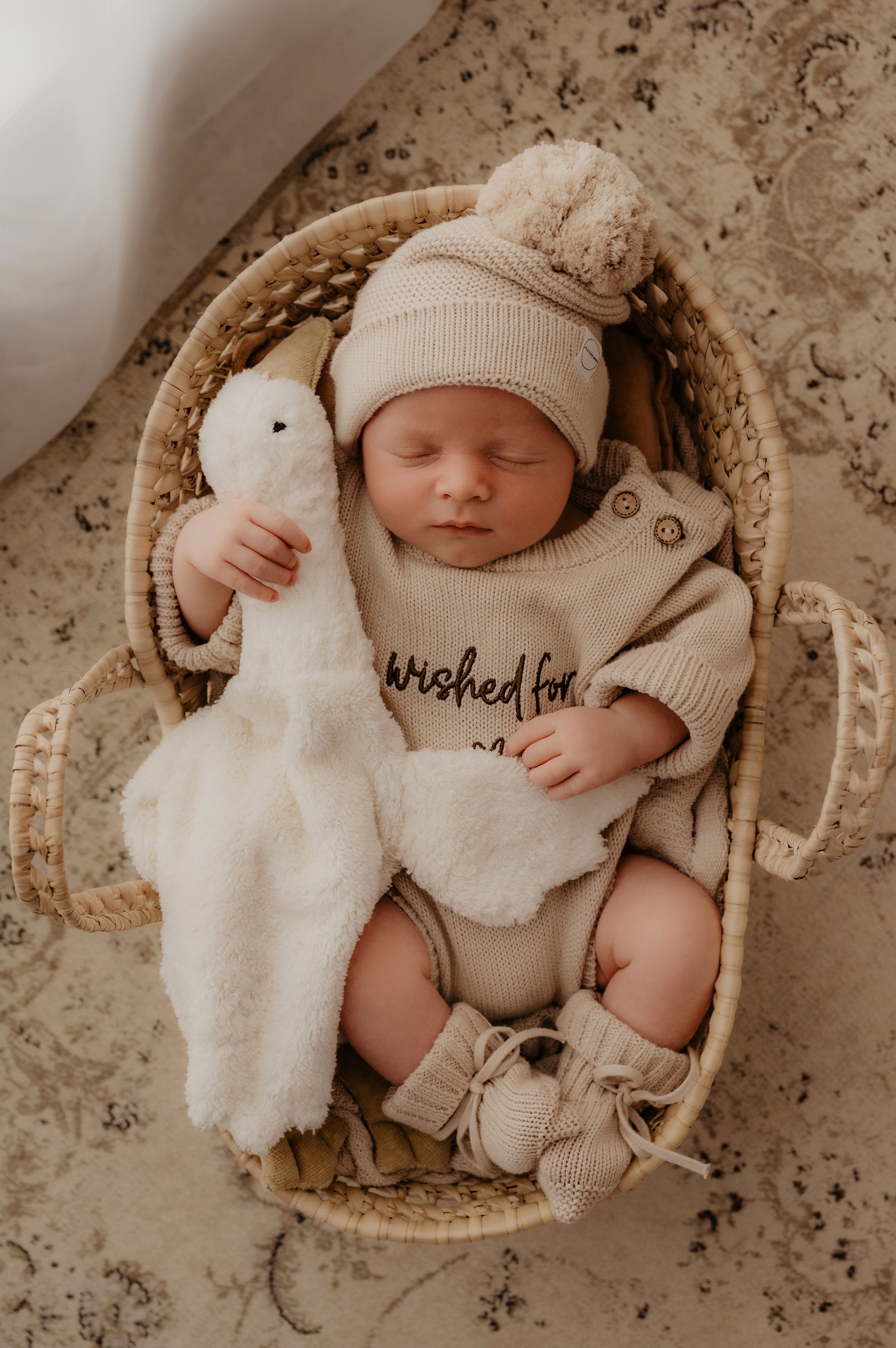 Newborn baby sleeping in cream-colored outfit with matching hat and plush bunny toy in rustic basket setting.
