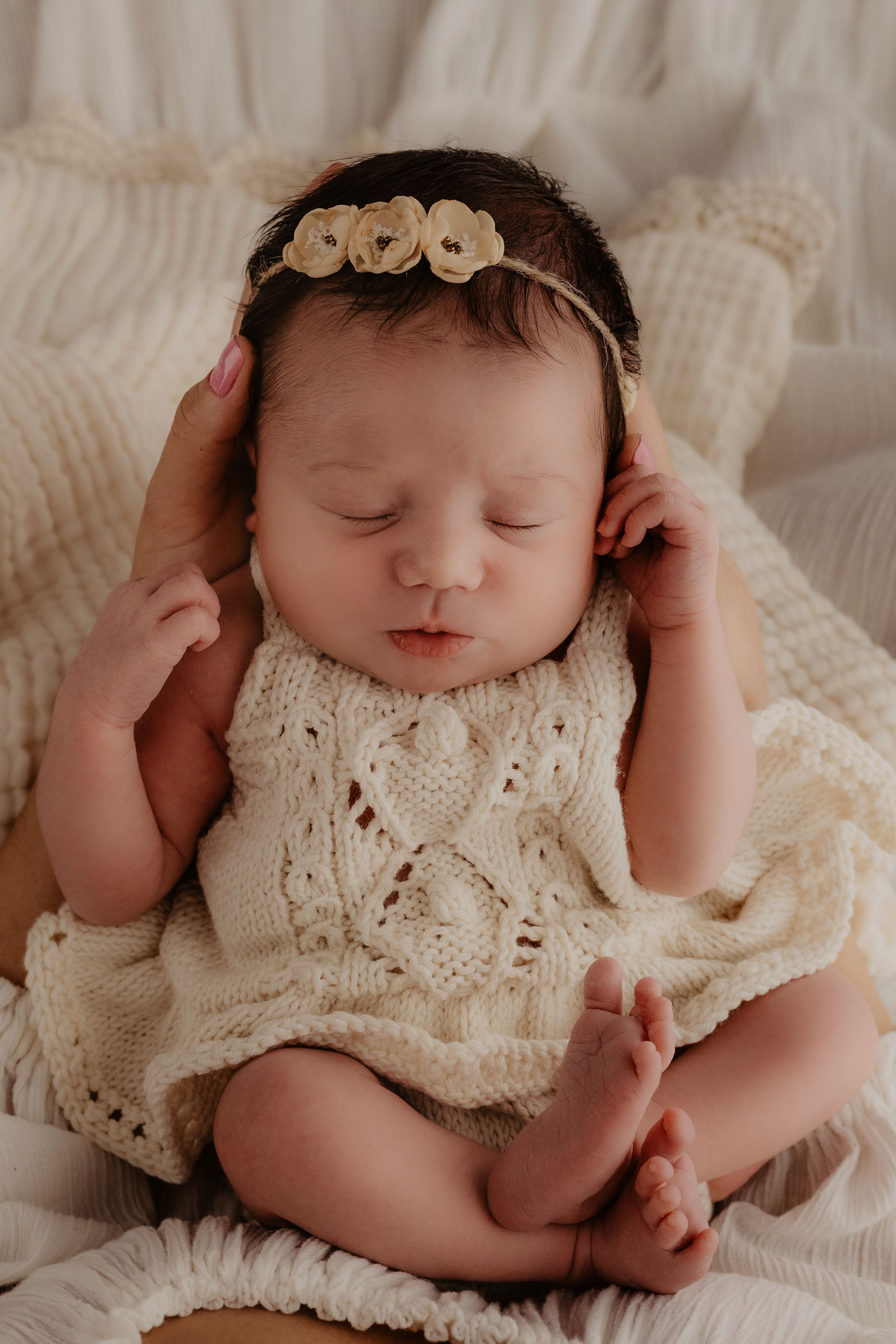 A sleeping newborn wearing a delicate cream-colored knit outfit lies on soft white fabric.