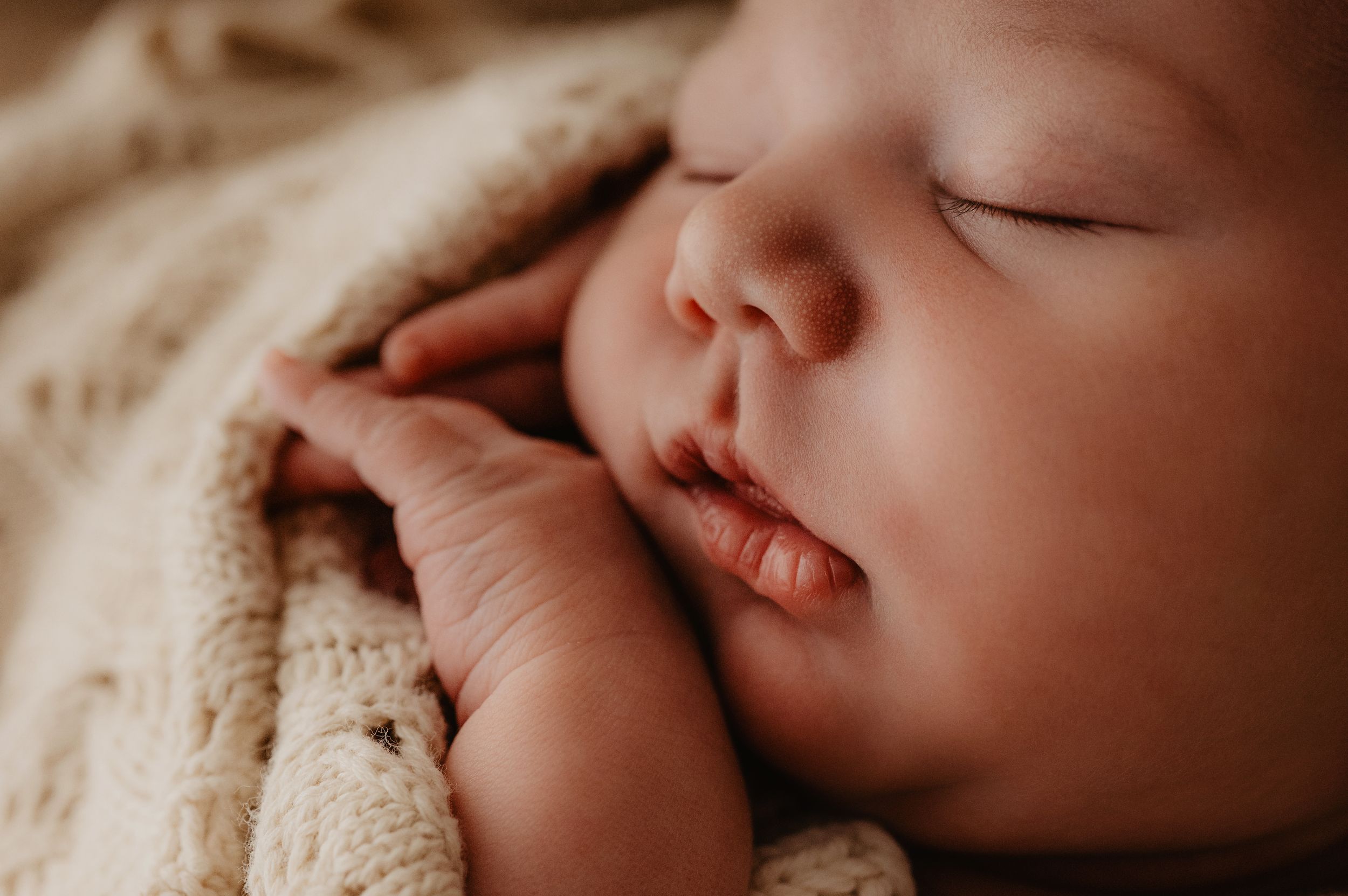 A sleeping baby wrapped in a cozy beige knit blanket lies peacefully against a soft textured background.