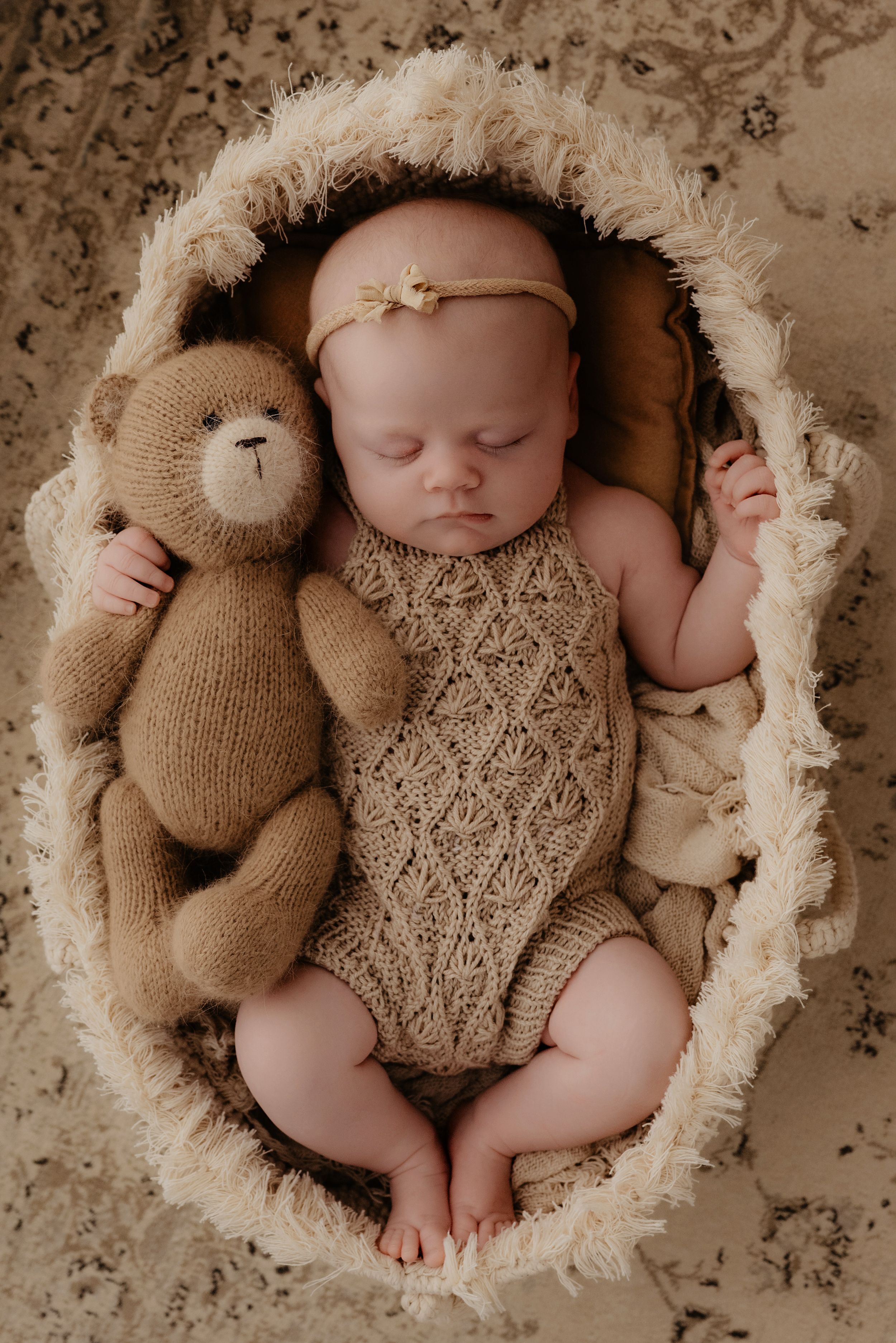 A newborn baby sleeping peacefully in a cream-colored basket with a knitted brown teddy bear and lace outfit.